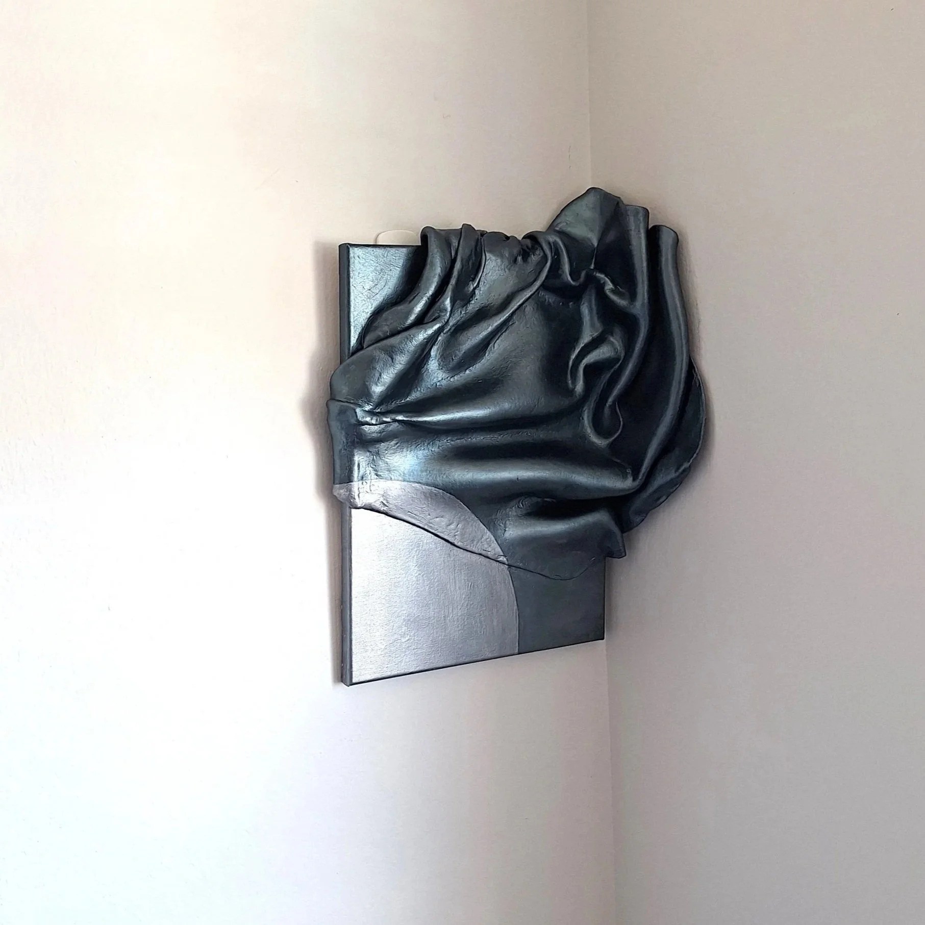 A black sculpture of a draped cloth on a gray rectangular base, displayed against a plain beige wall.