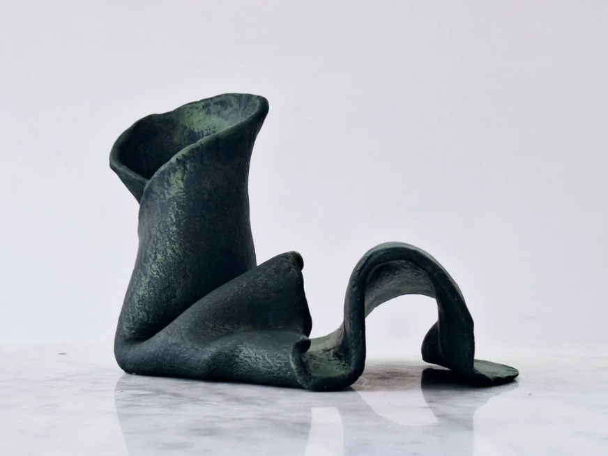 A dark, abstract sculpture with a curving, ribbon-like shape set against a plain white background.