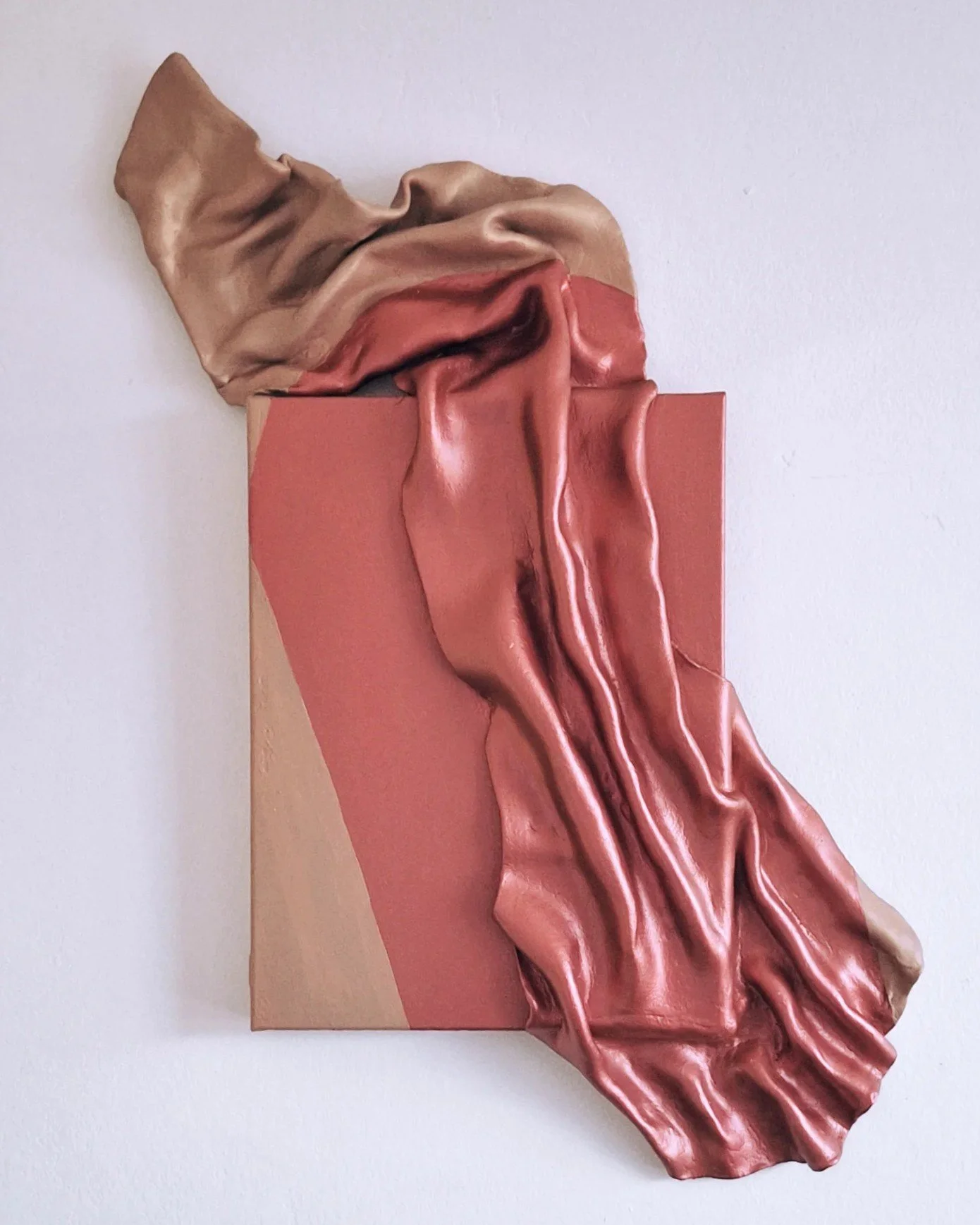 A mixed media art piece featuring a textured, layered sculpture with metallic and matte finish in earth tones, resembling a flowing, abstract form mounted on a flat background.