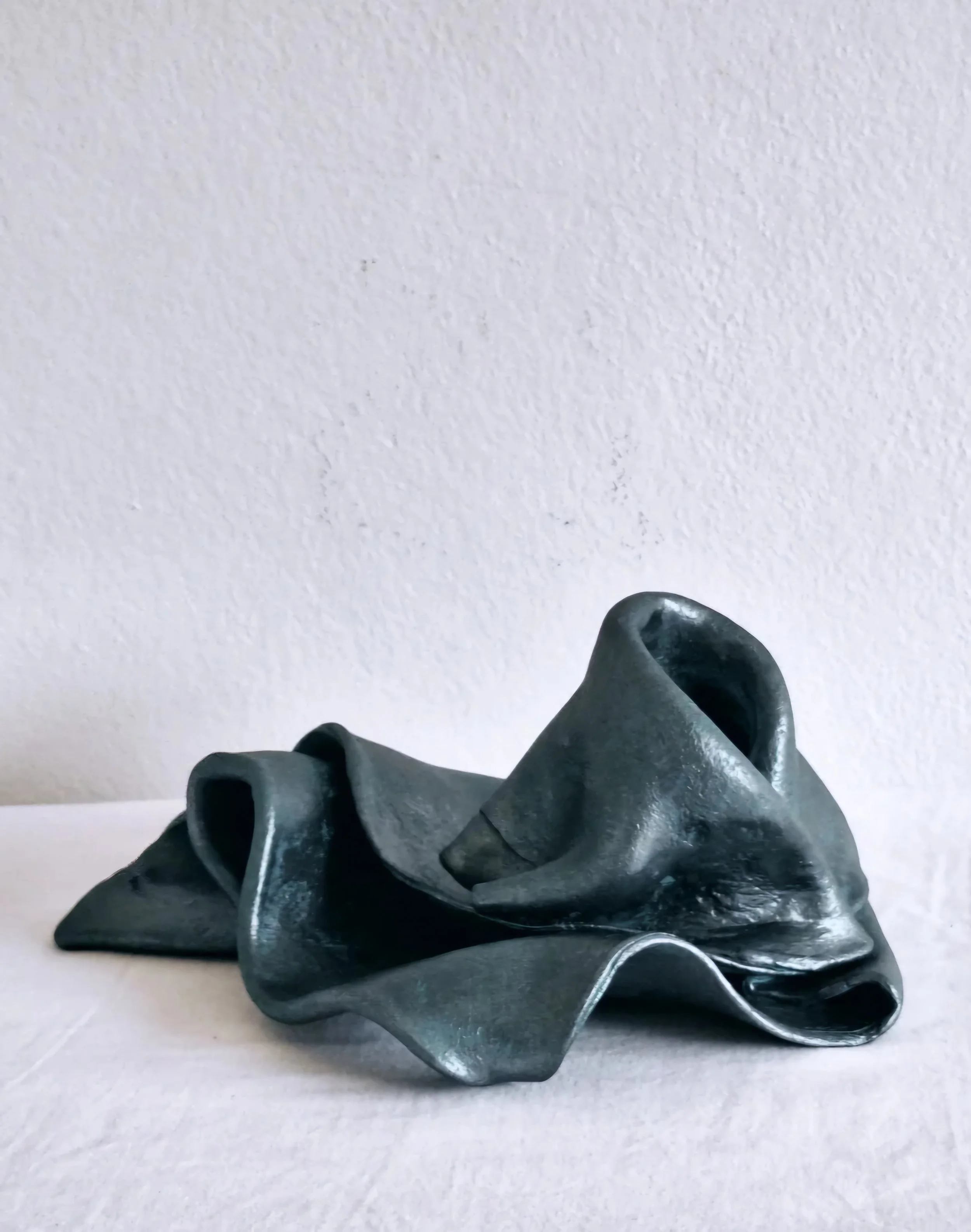 A strange silvery-blue twisted vessel resting on a white, textured surface, with a light-colored wall in the background.