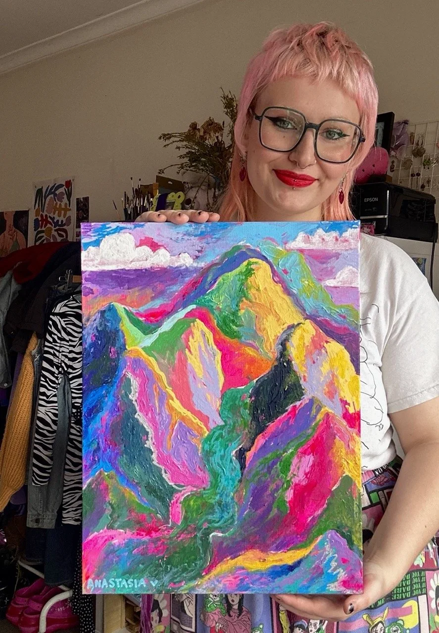 A woman with pink hair, glasses, and red lipstick holding a colorful painting of a mountain landscape with vivid neon colors including pink, green, yellow, and purple.