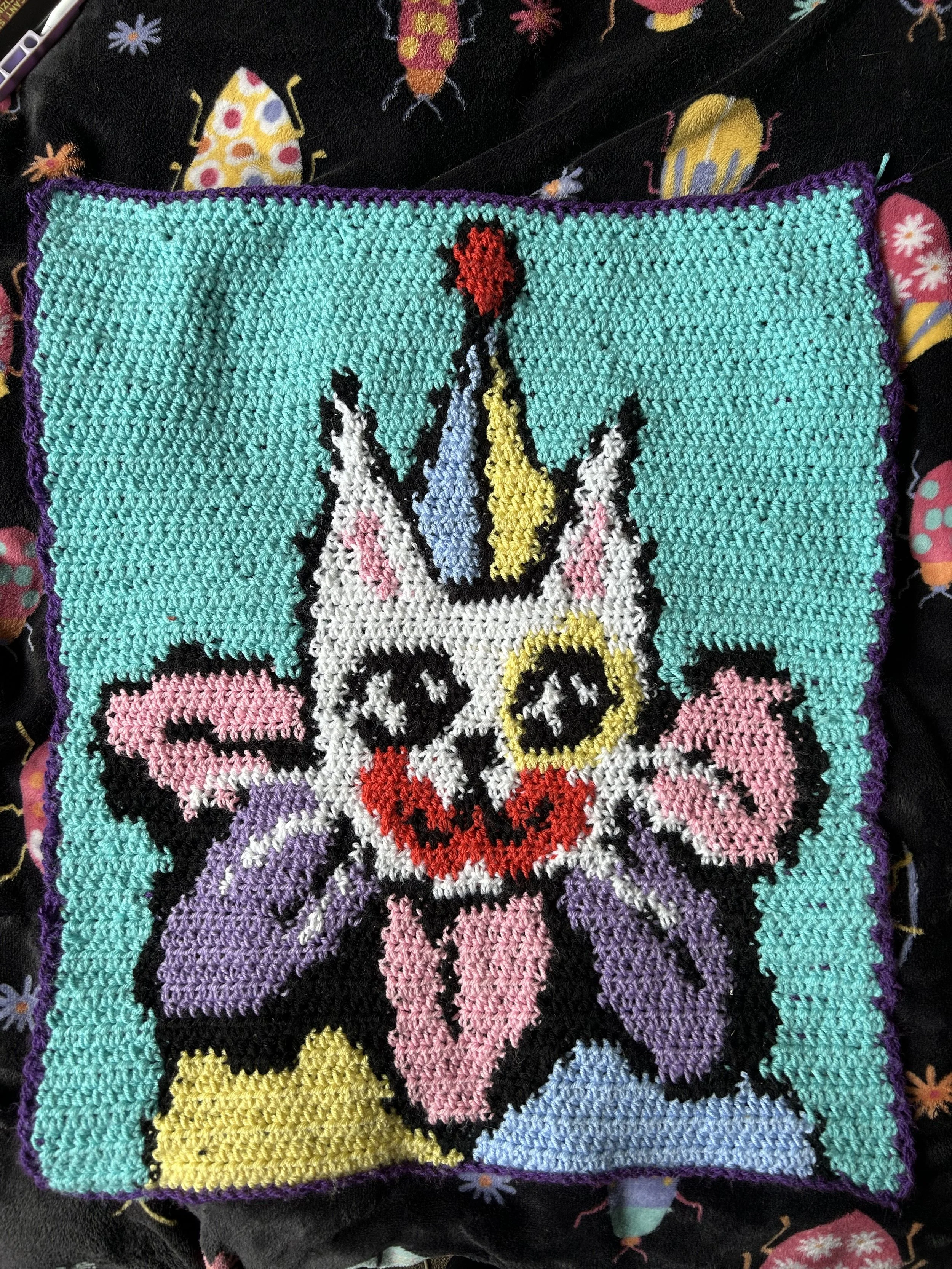 Colorful cross-stitch blanket featuring a unicorn with a rainbow-colored horn, pink cheeks, and a yellow mane, set against a bright turquoise background with a purple border.