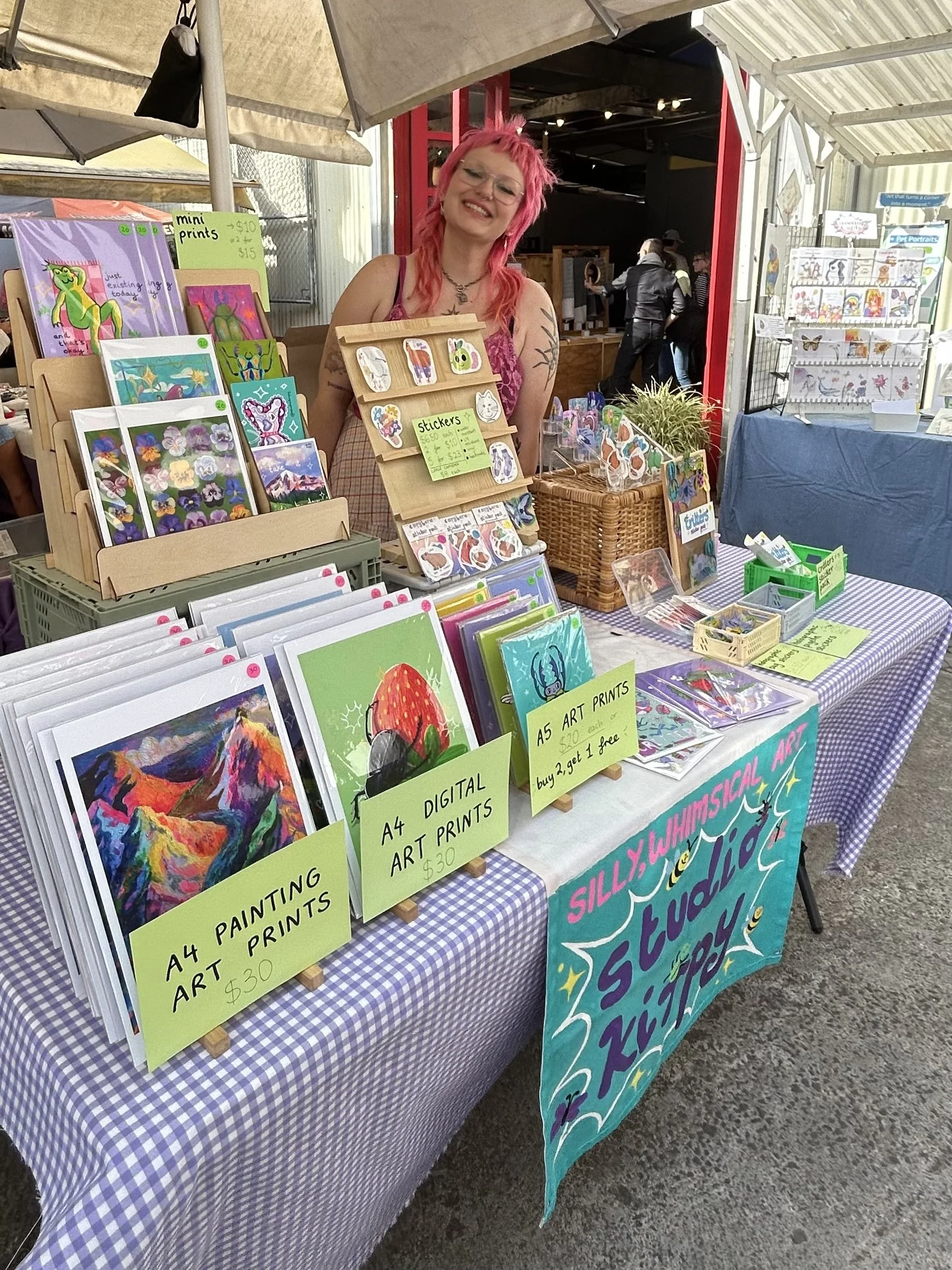A woman with pink hair standing behind a table at an outdoor art and craft fair, displaying colorful paintings, stickers, and prints for sale.