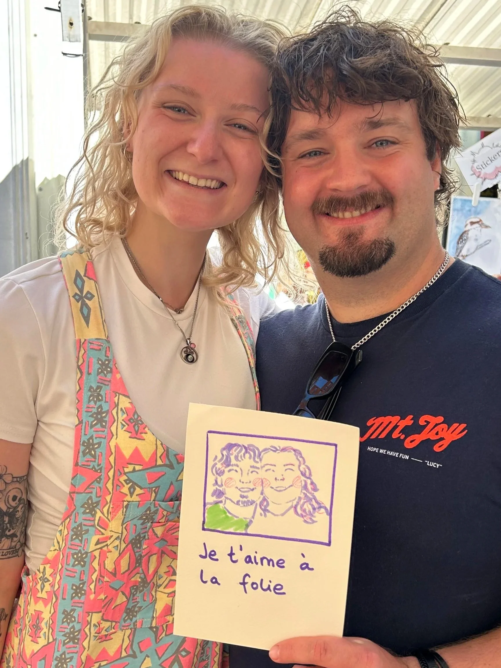 A smiling woman with curly blonde hair standing next to a smiling man with curly dark hair, both taking a selfie outdoors. The woman has a colorful strap over her shoulder and is wearing a necklace. The man is wearing a dark T-shirt with red and white text, and sunglasses hanging from his collar. They are holding a drawing with a message that says, 'Je t'aime à la folie' in French, featuring a colorful sketch of smiling faces.