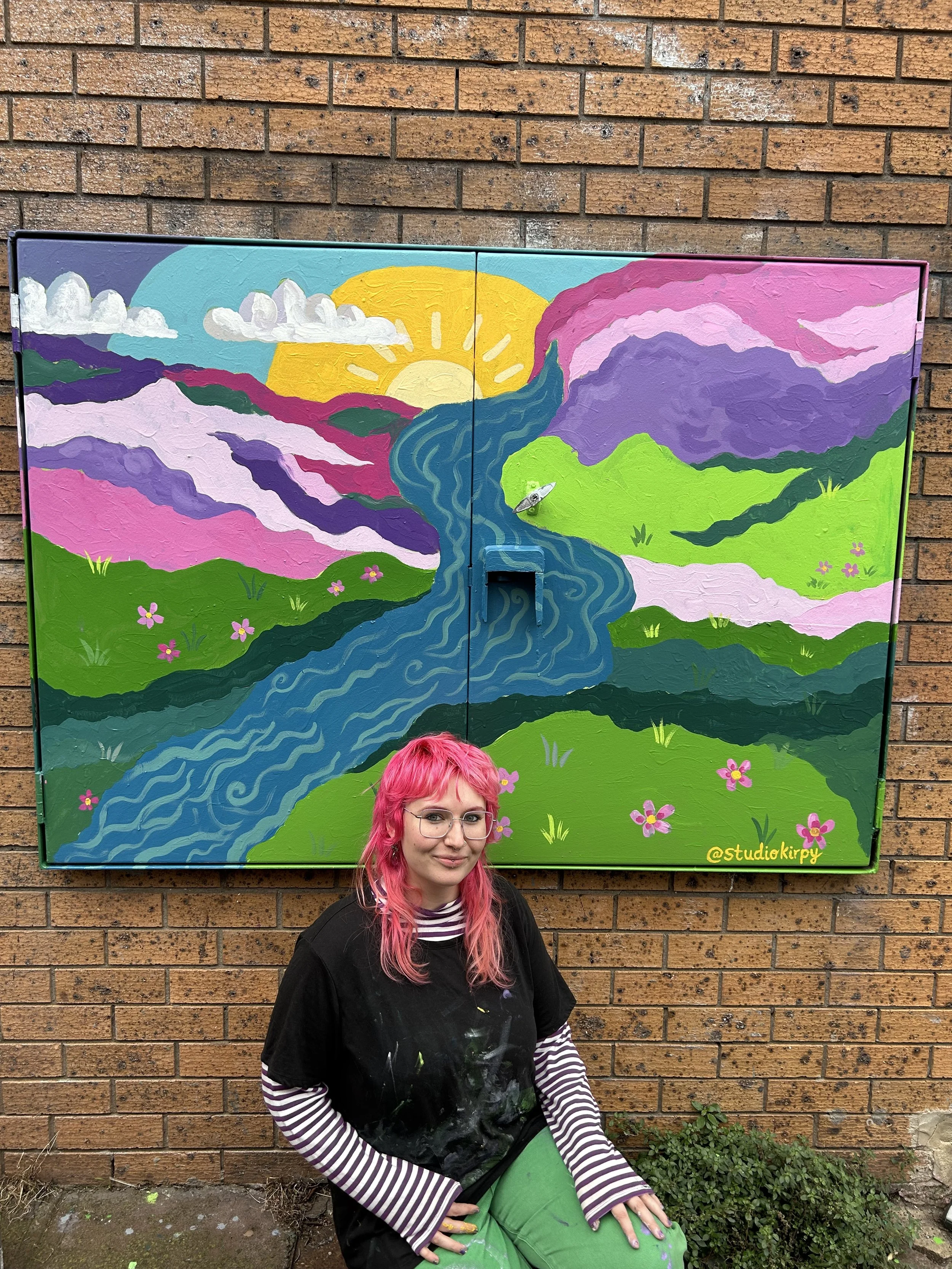 Young woman with pink hair and glasses sitting against a brick wall, wearing a black apron over a striped long-sleeve shirt and green pants, in front of a colorful mural painting of a sunrise over a landscape with mountains, river, and flowers.