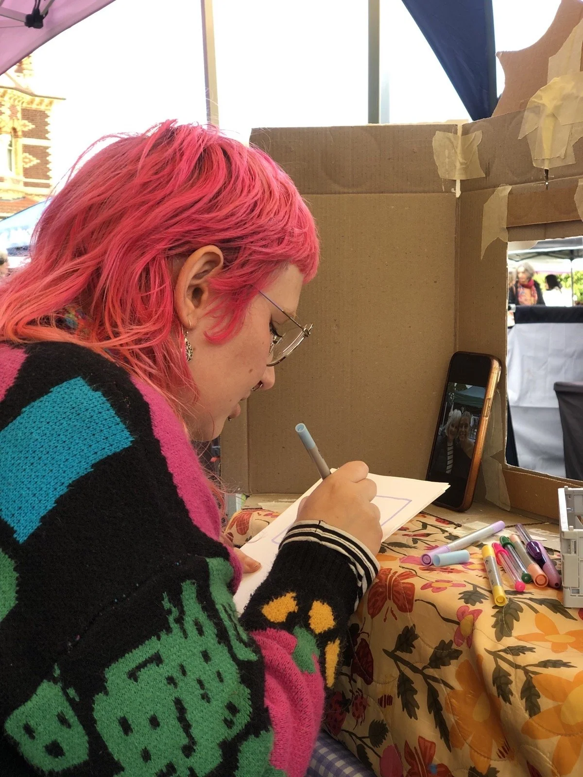 A woman with pink hair drawing on paper at a booth, with a smartphone displaying her image, markers on a table, and some people visible in the background.