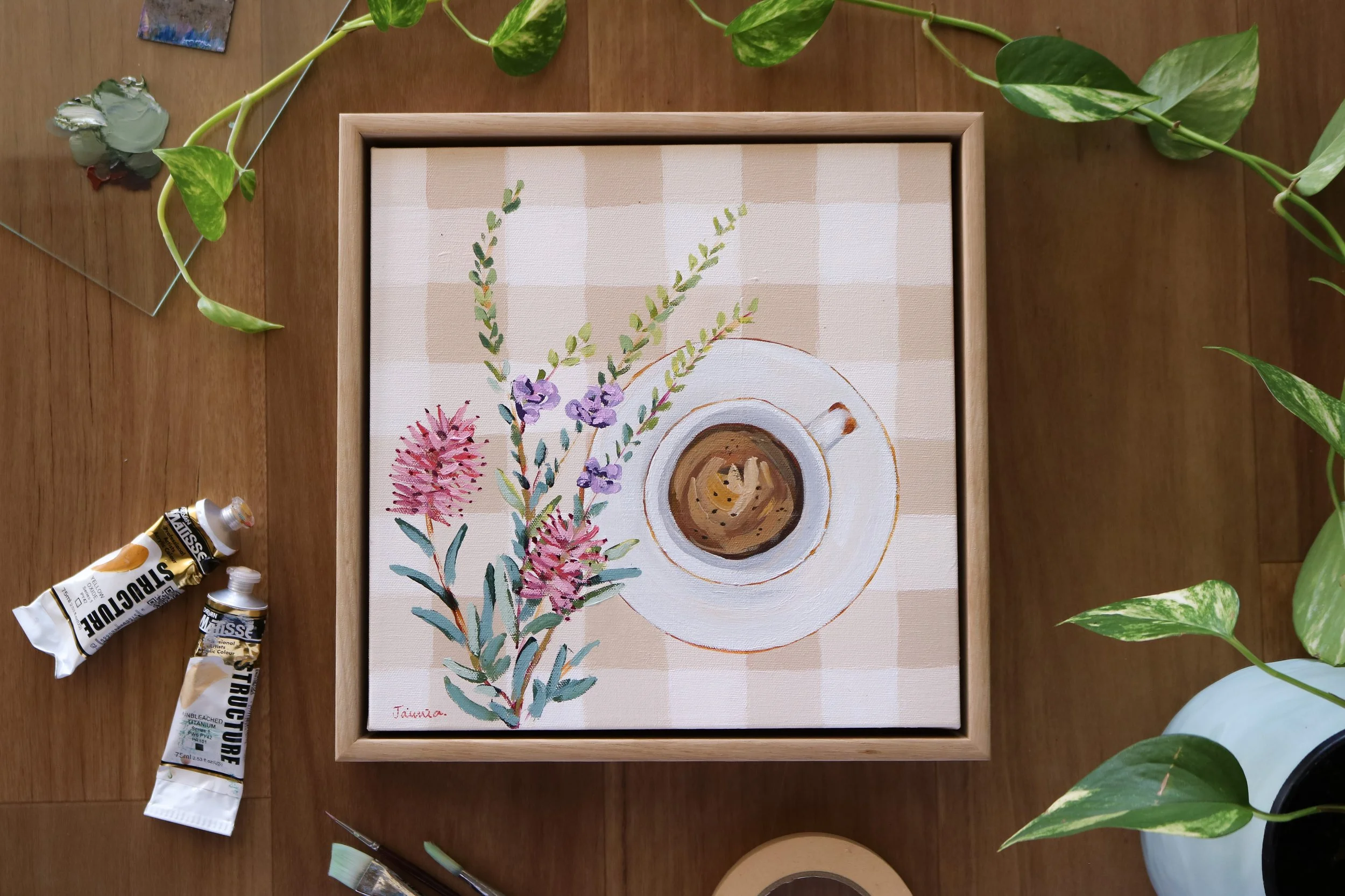 A painting of flowers and a coffee cup on a checkered tablecloth. The painting is inside a wooden frame and is surrounded by plants, paint tubes, and art supplies.