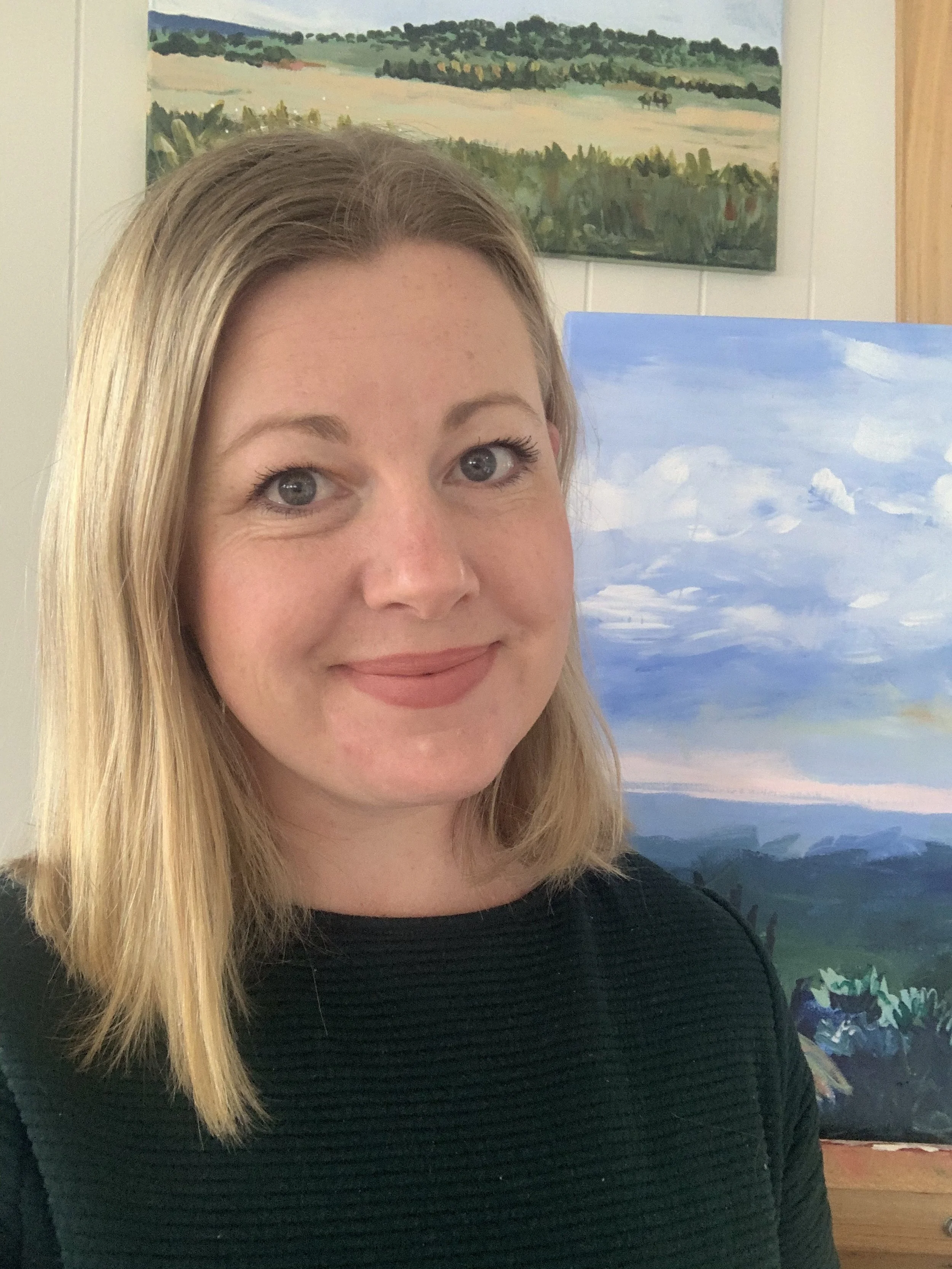 A woman with shoulder-length blonde hair and blue eyes smiling at the camera. She is wearing a black top and standing in front of framed landscape paintings, one of which depicts a cloudy sky.
