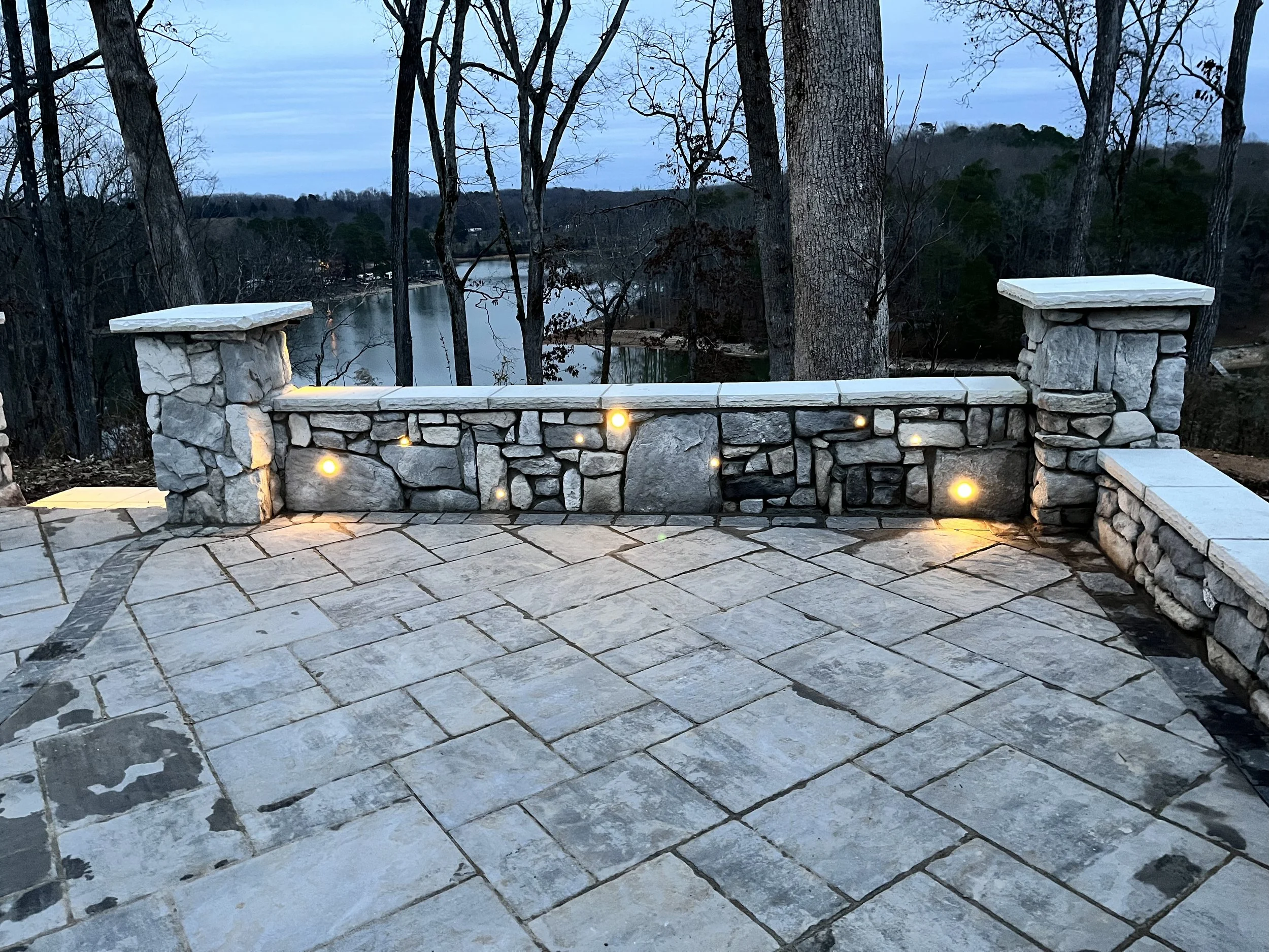 Nighttime view of a wooden and stone fence with small lights, overlooking a river surrounded by trees with mostly bare branches and a distant forested hill.