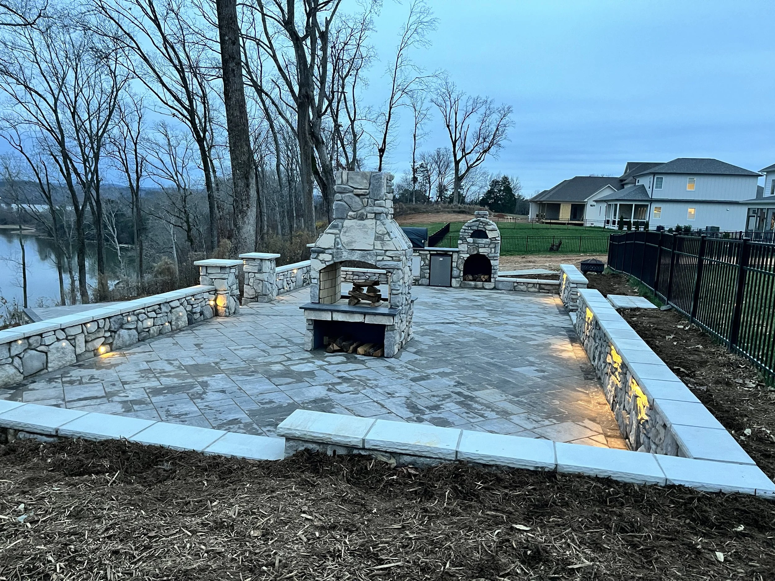 A backyard patio area with stone fireplace and outdoor kitchen, surrounded by a black metal fence, with trees and a house in the background during dusk.