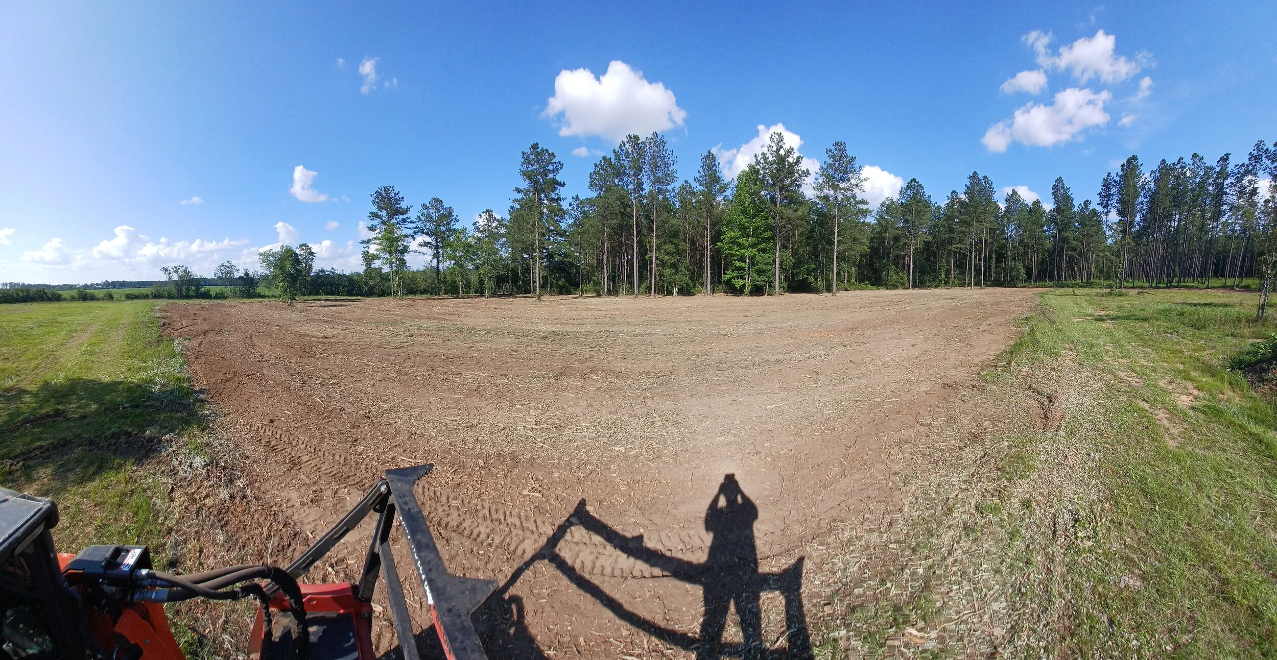 A wide open field with bare soil and a line of tall pine trees in the background. The sky is bright blue with scattered white clouds. A shadow of a person taking a photo and part of a tractor or construction vehicle is visible in the foreground.