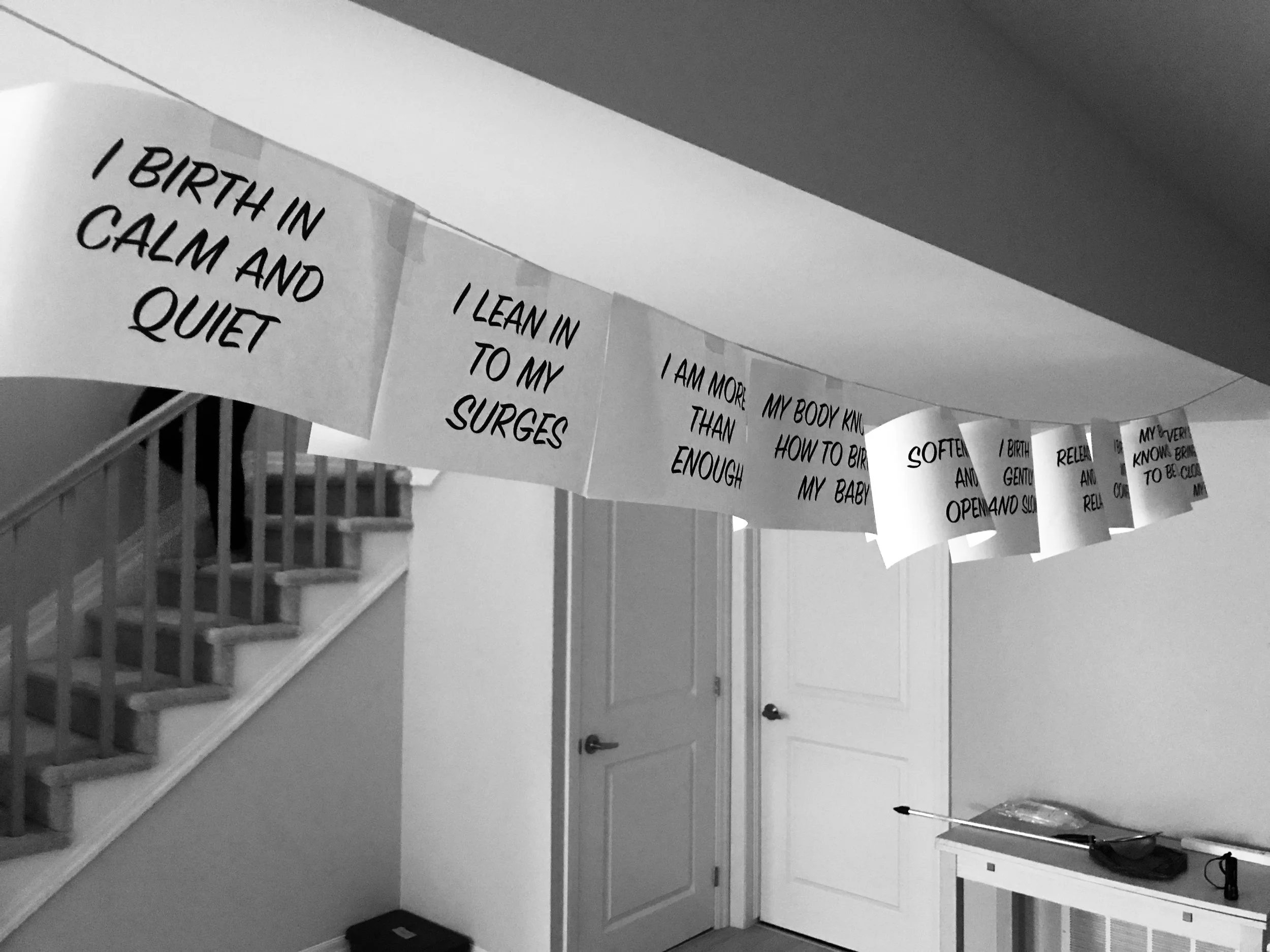 Paper notes with handwritten affirmations hanging on a string in a home interior, near a staircase and a closed door.