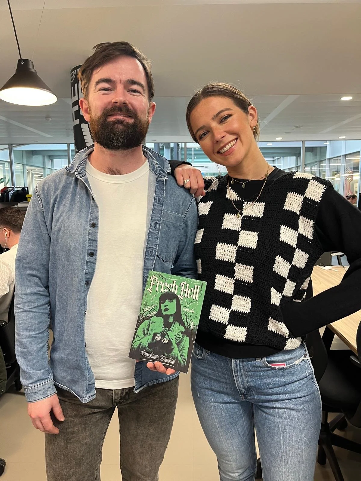 A man with a beard and a woman with short hair pose together in a modern indoor setting. The man is holding a comic book titled 'Flesh Hell'. The woman is wearing a black and white checkered sweater. Nicole Vanner and Andrew Hadley
