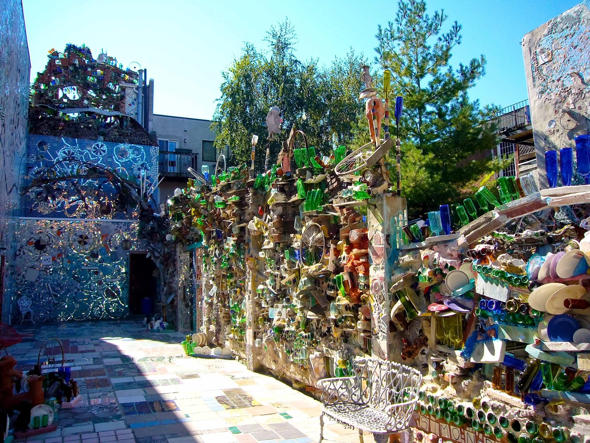 A vibrant, immersive outdoor mosaic art installation by Isaiah Zagar. The narrow walkway is surrounded by high walls completely covered in intricate folk-art mosaics