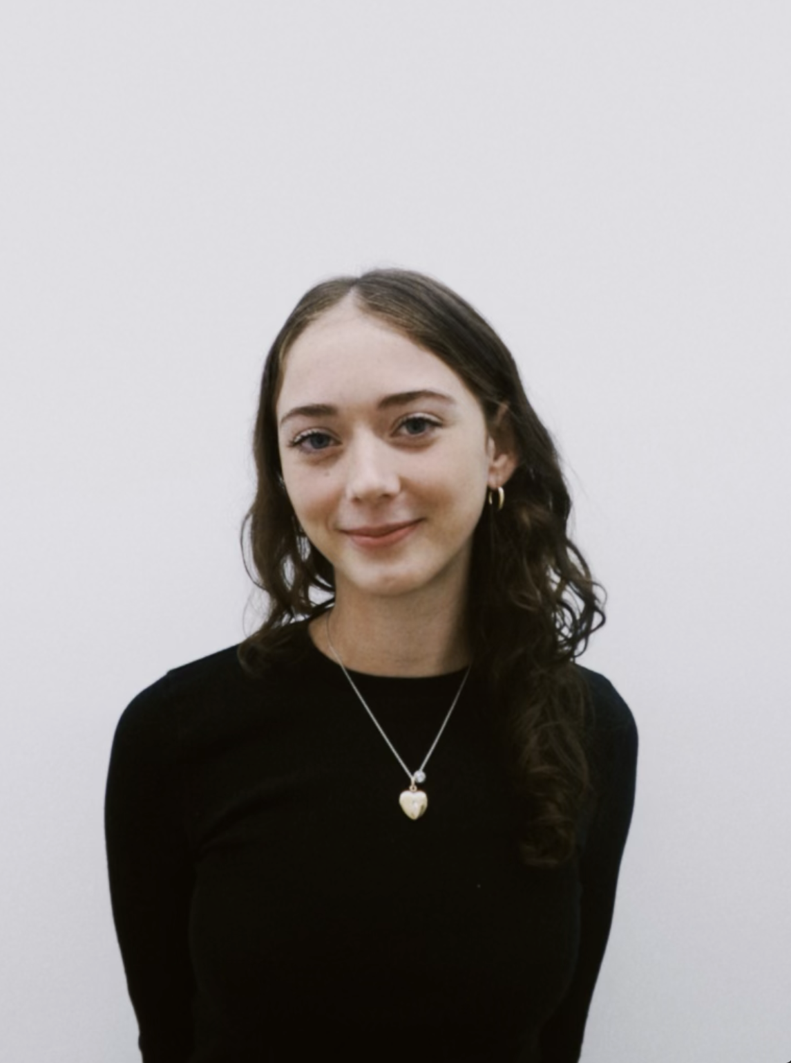 Portrait of a young woman with brown, wavy hair, wearing a black top, hoop earrings, and a necklace with a heart-shaped pendant, standing against a plain white background.