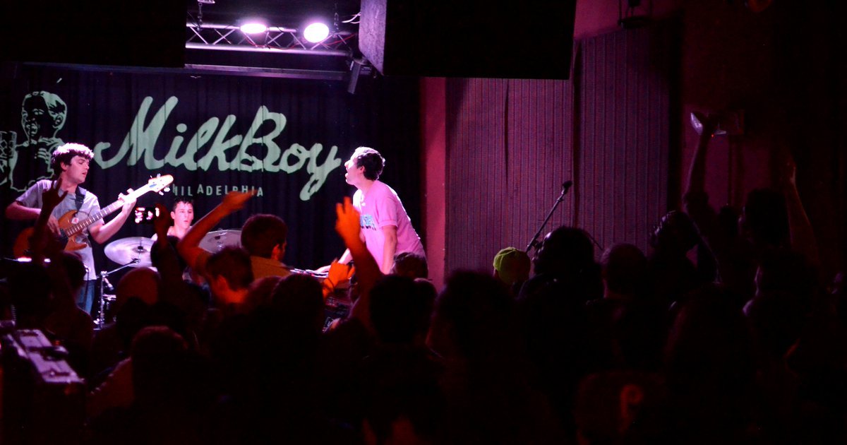 A wide-angle shot of a live concert at MilkBoy Philly. A band is performing on a small, elevated stage illuminated by intense magenta and blue stage lights. The lead singer is mid-performance, holding a microphone.