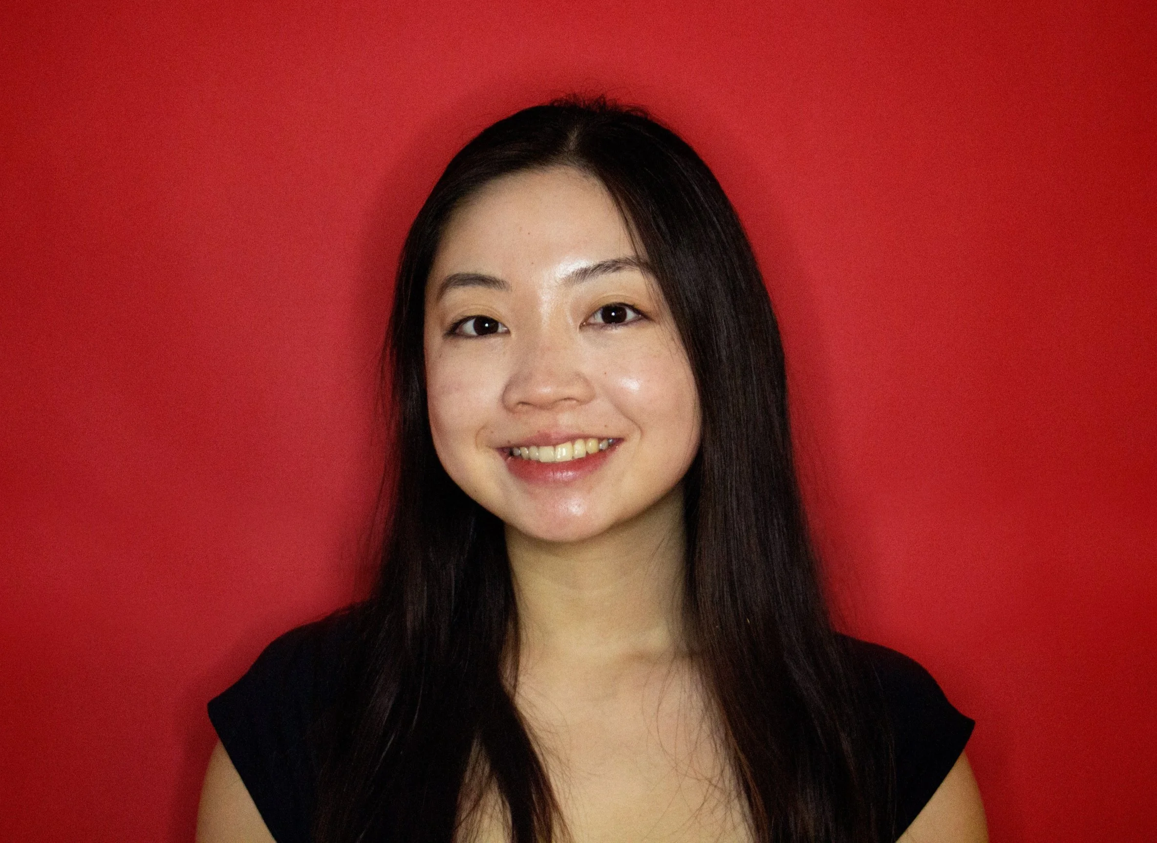Portrait of a young woman with long dark hair smiling in front of a red background.