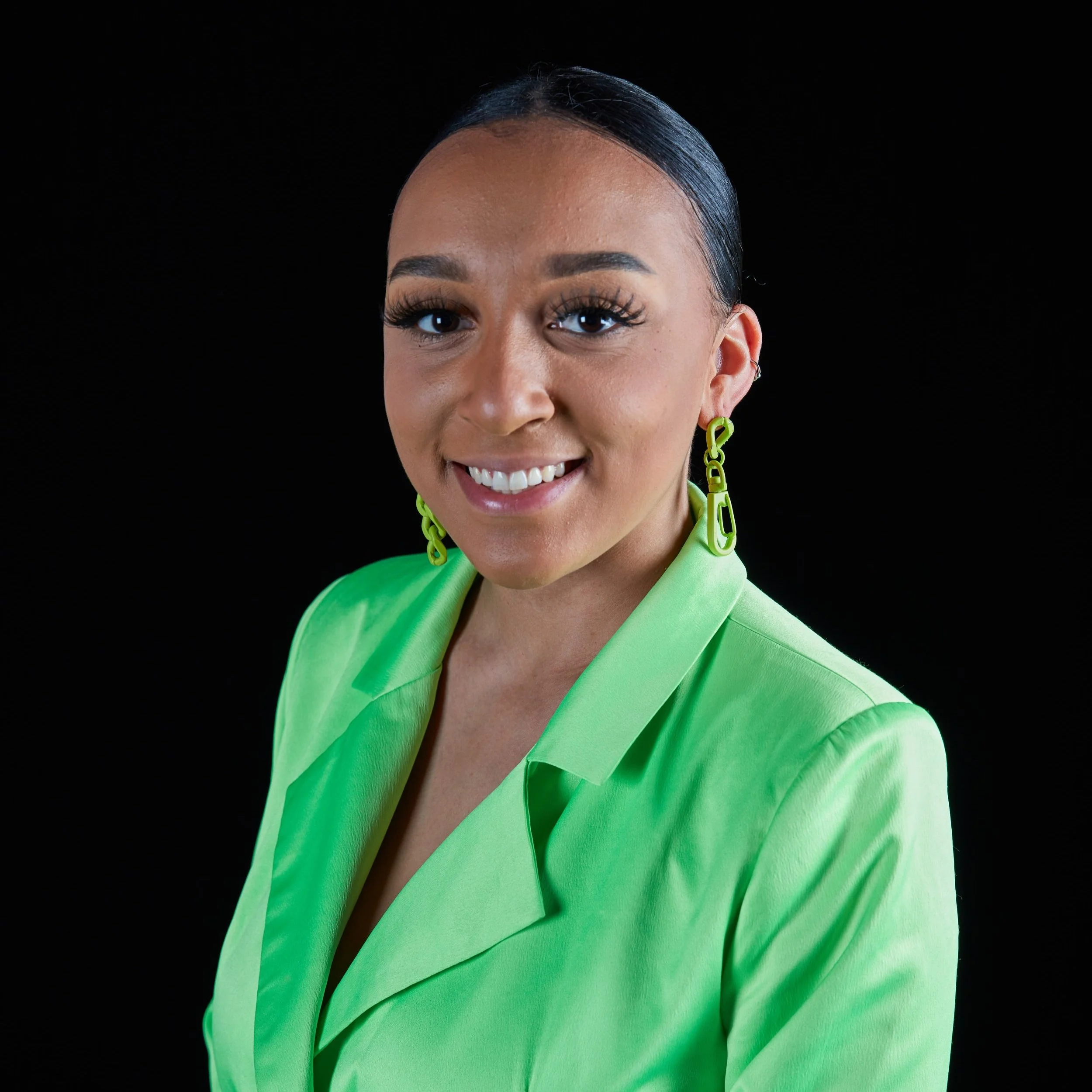 A woman smiling, wearing a bright green blazer and matching earrings, against a black background.