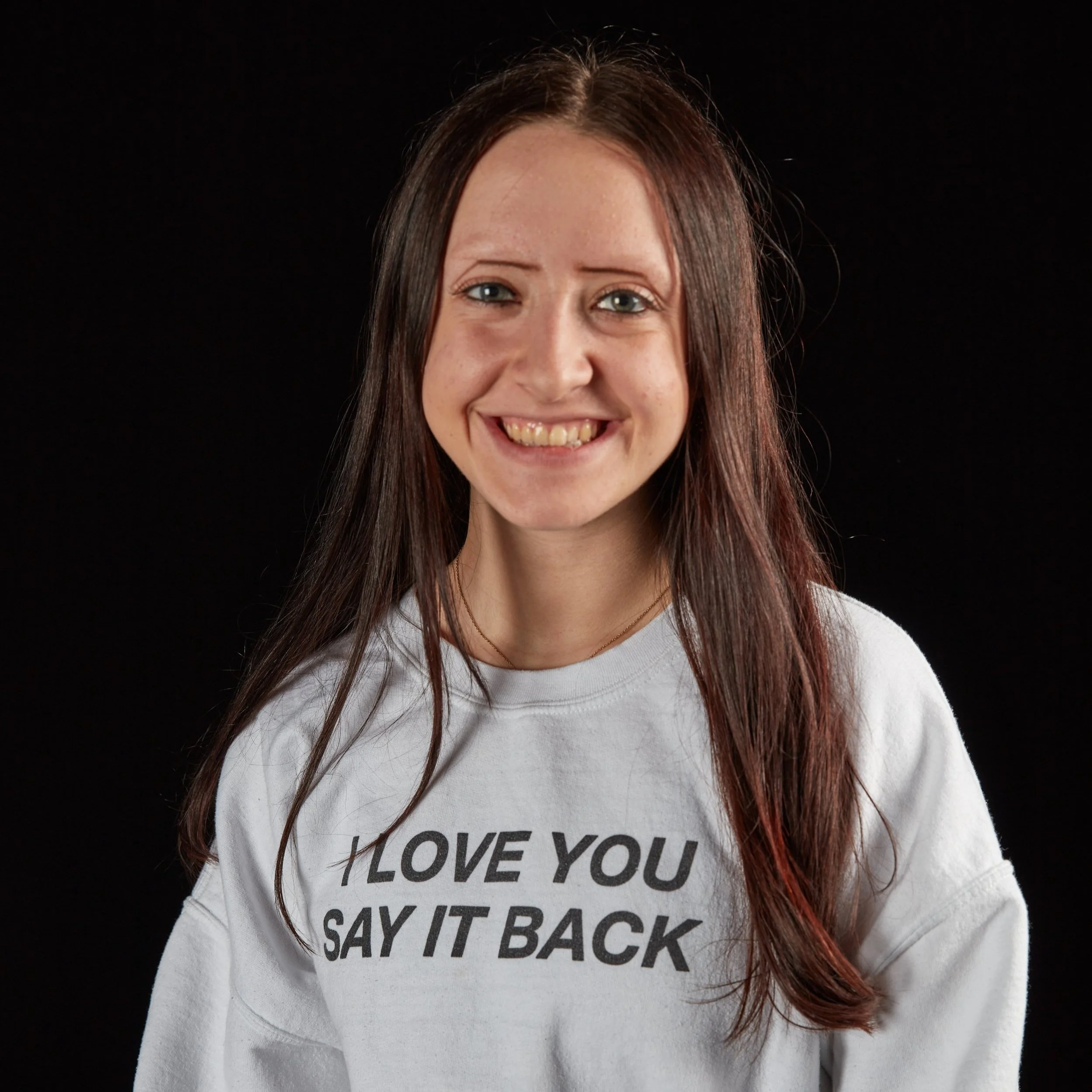 Smiling woman with long brown hair wearing a white sweatshirt that says 'I LOVE YOU SAY IT BACK' against a black background.