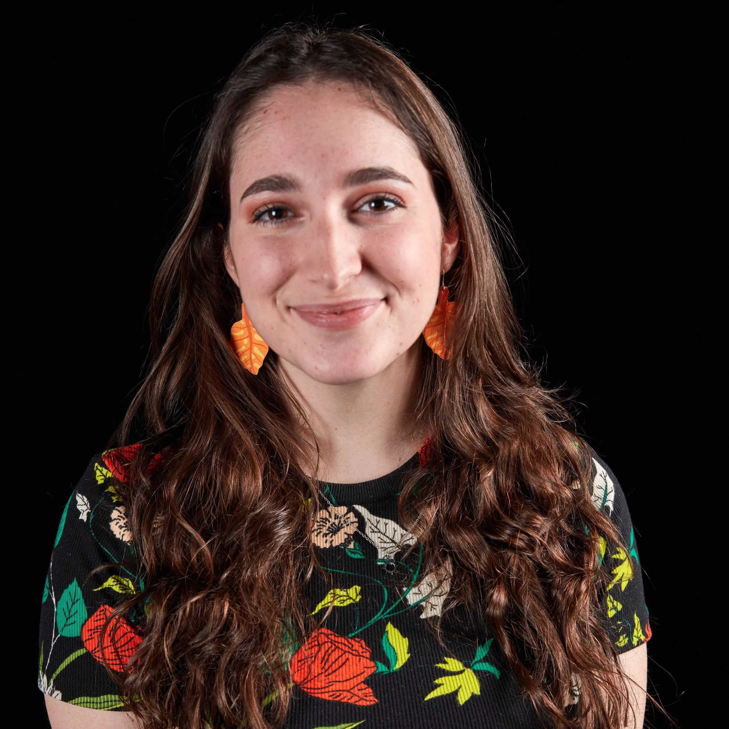 A young woman with curly brown hair and light skin, smiling, wearing orange leaf-shaped earrings and a black floral top against a black background.