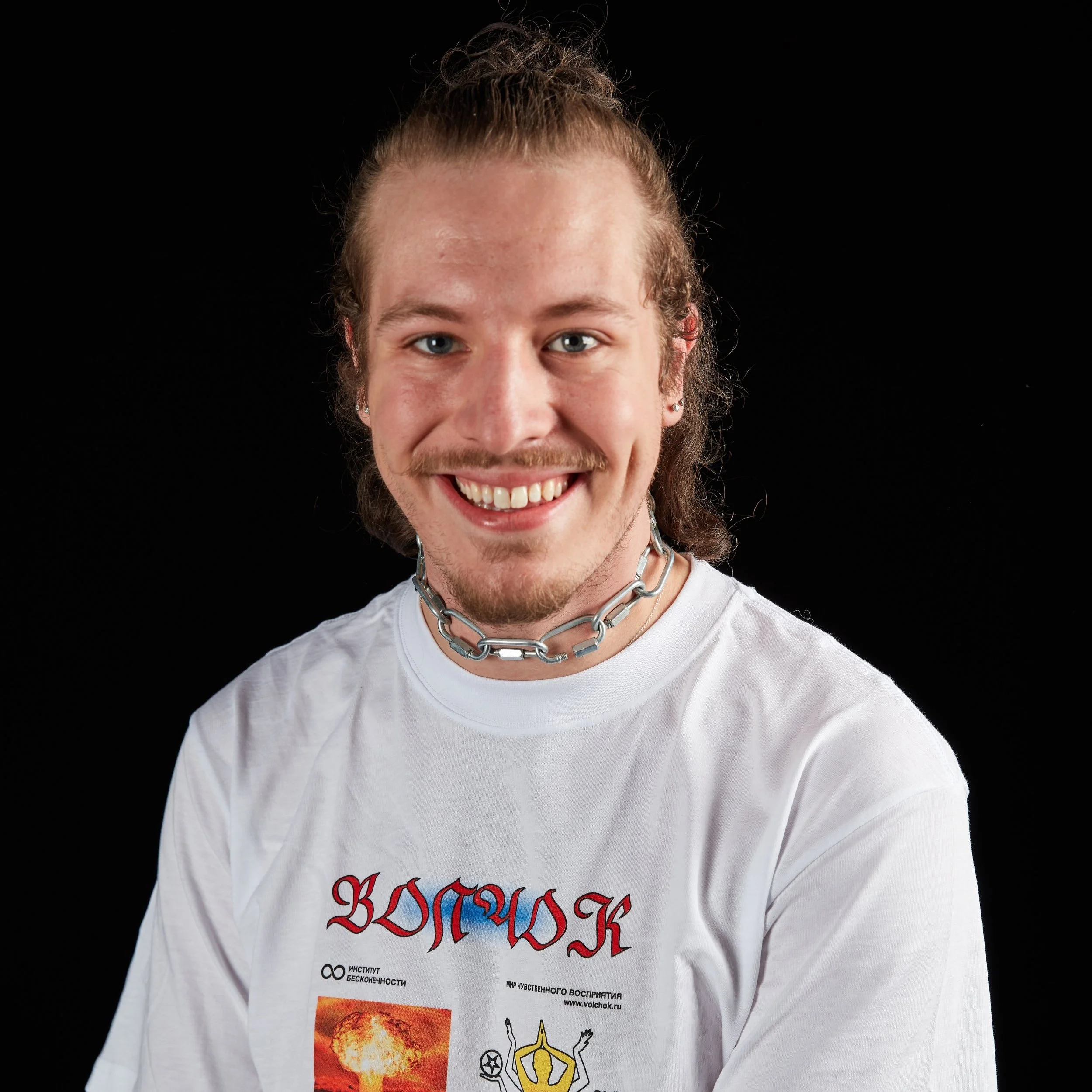 Young man with long hair in a bun, wearing a white T-shirt with Russian text and graphics, and a metal chain necklace, smiling against a black background.