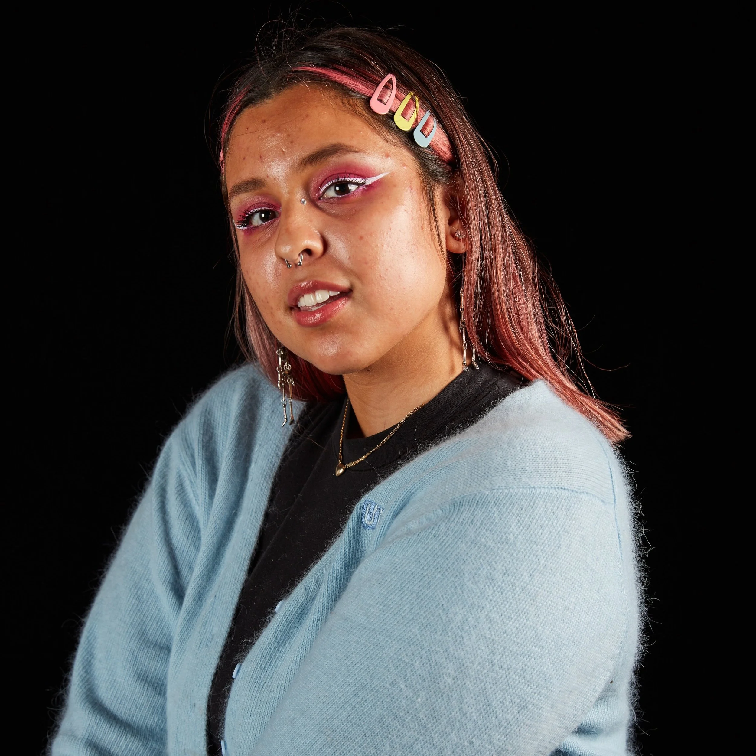 A young woman with pink highlights in her hair, wearing hair clips, makeup with pink eyeshadow, winged eyeliner, and jewelry, dressed in a black shirt and a light blue cardigan, posing against a black background.