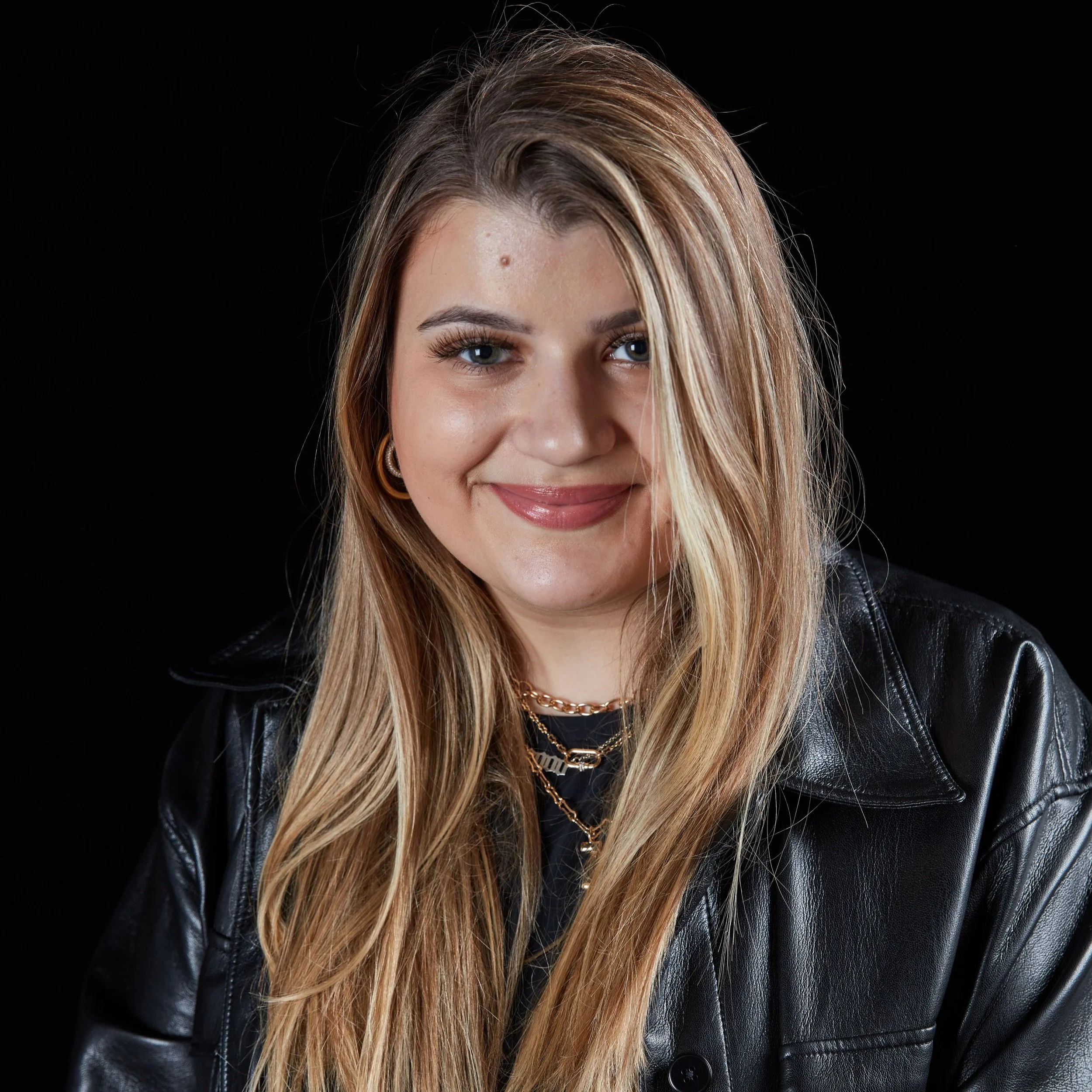 A young woman with long blonde hair, wearing a black leather jacket and gold necklaces, smiling against a black background.