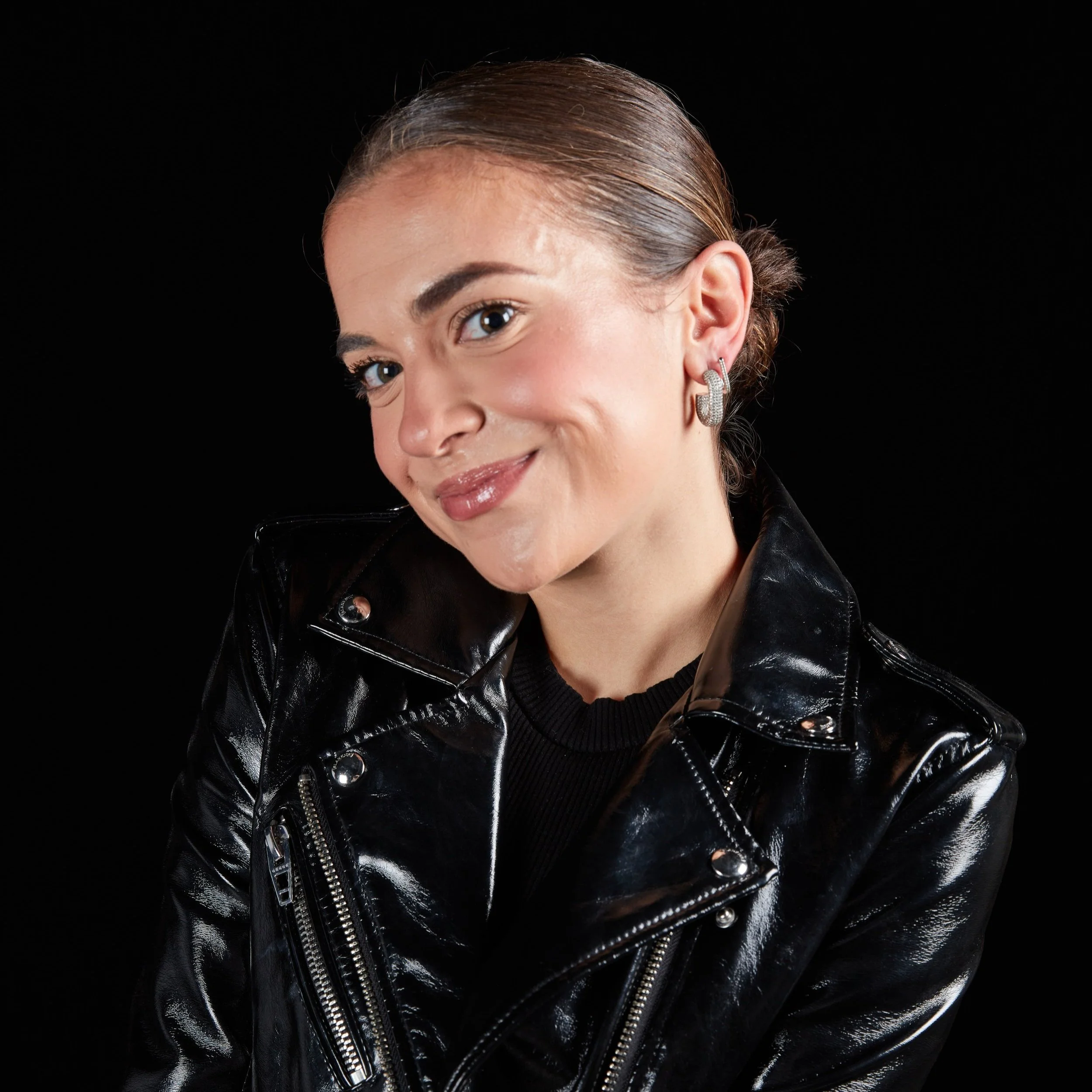 Young woman with slicked-back hair, wearing a black leather jacket and silver hoop earrings, smiling against a black background.