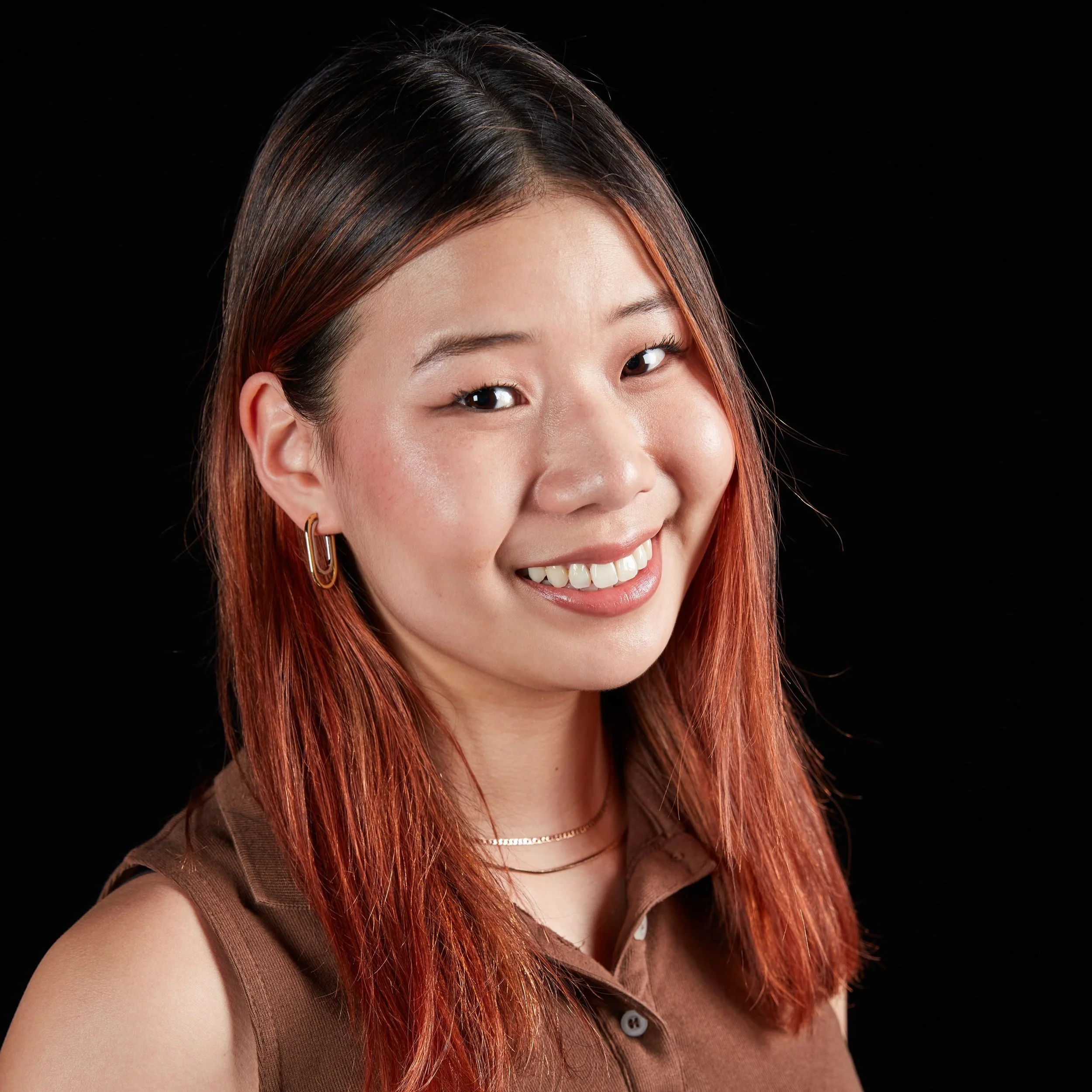 Portrait of a smiling young woman with reddish-brown hair, wearing a brown sleeveless top, gold earrings, and layered necklaces, against a black background.