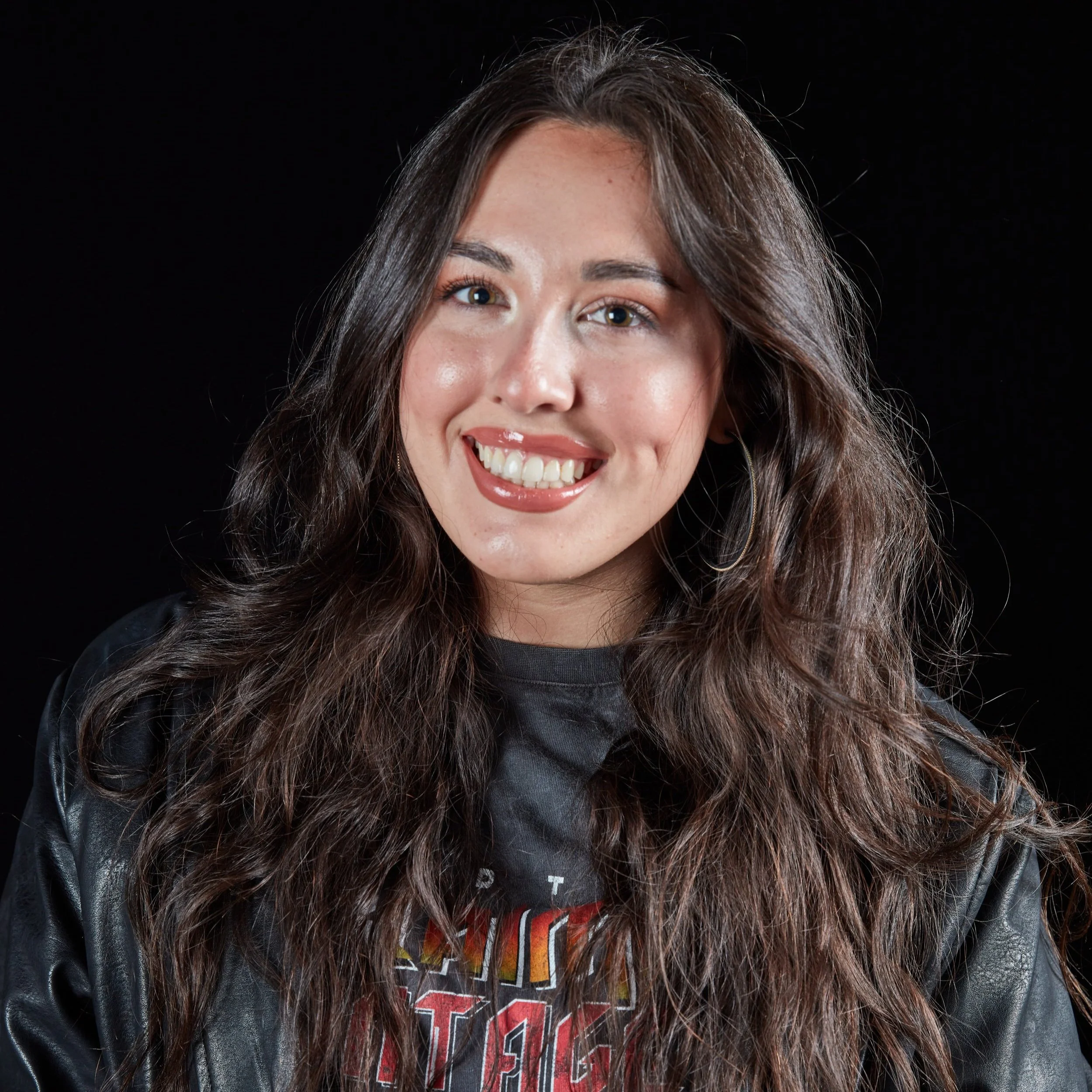 A young woman with long, wavy brown hair, wearing a black leather jacket and a graphic T-shirt, smiling against a black background.