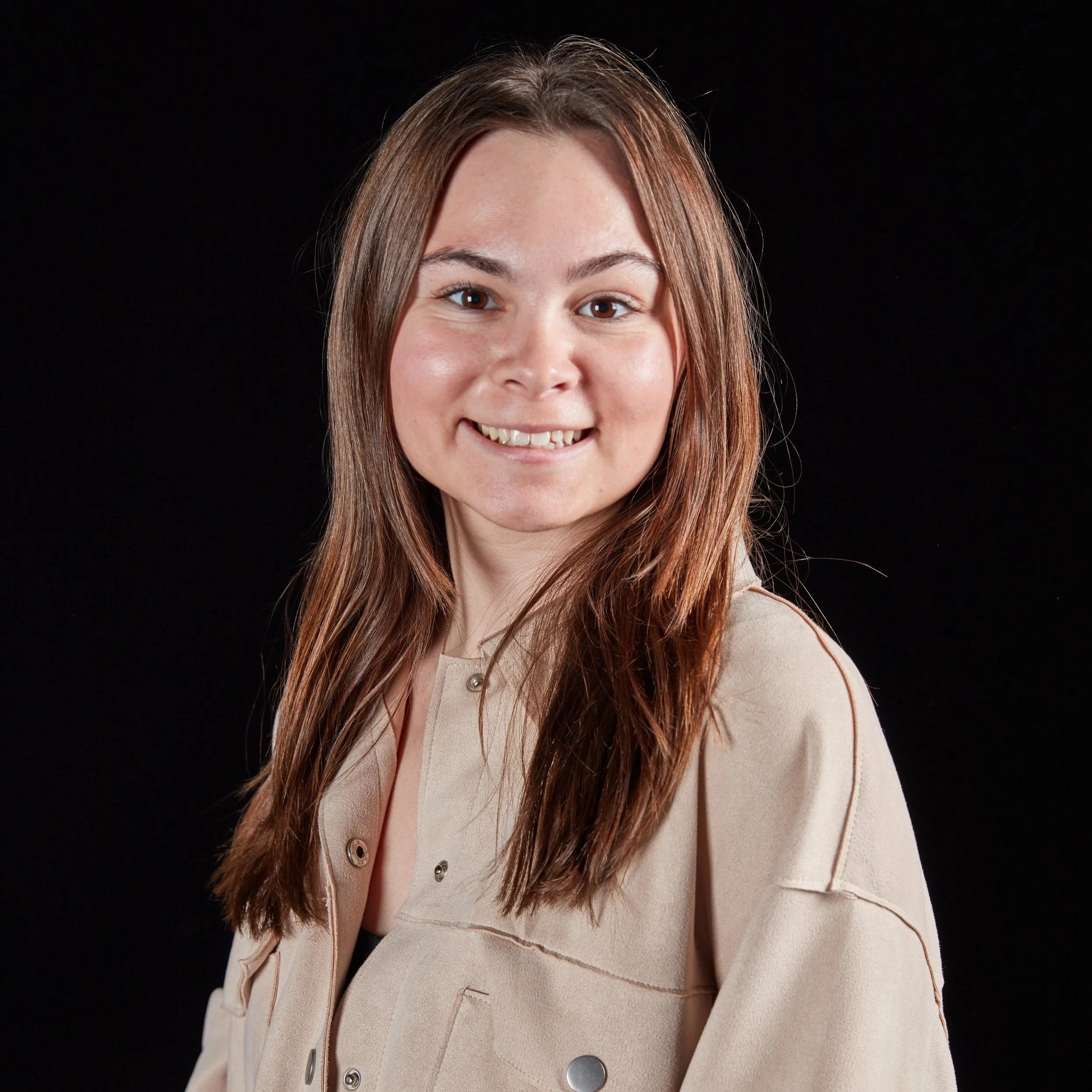 Portrait of a young woman with long brown hair, smiling, against a black background.