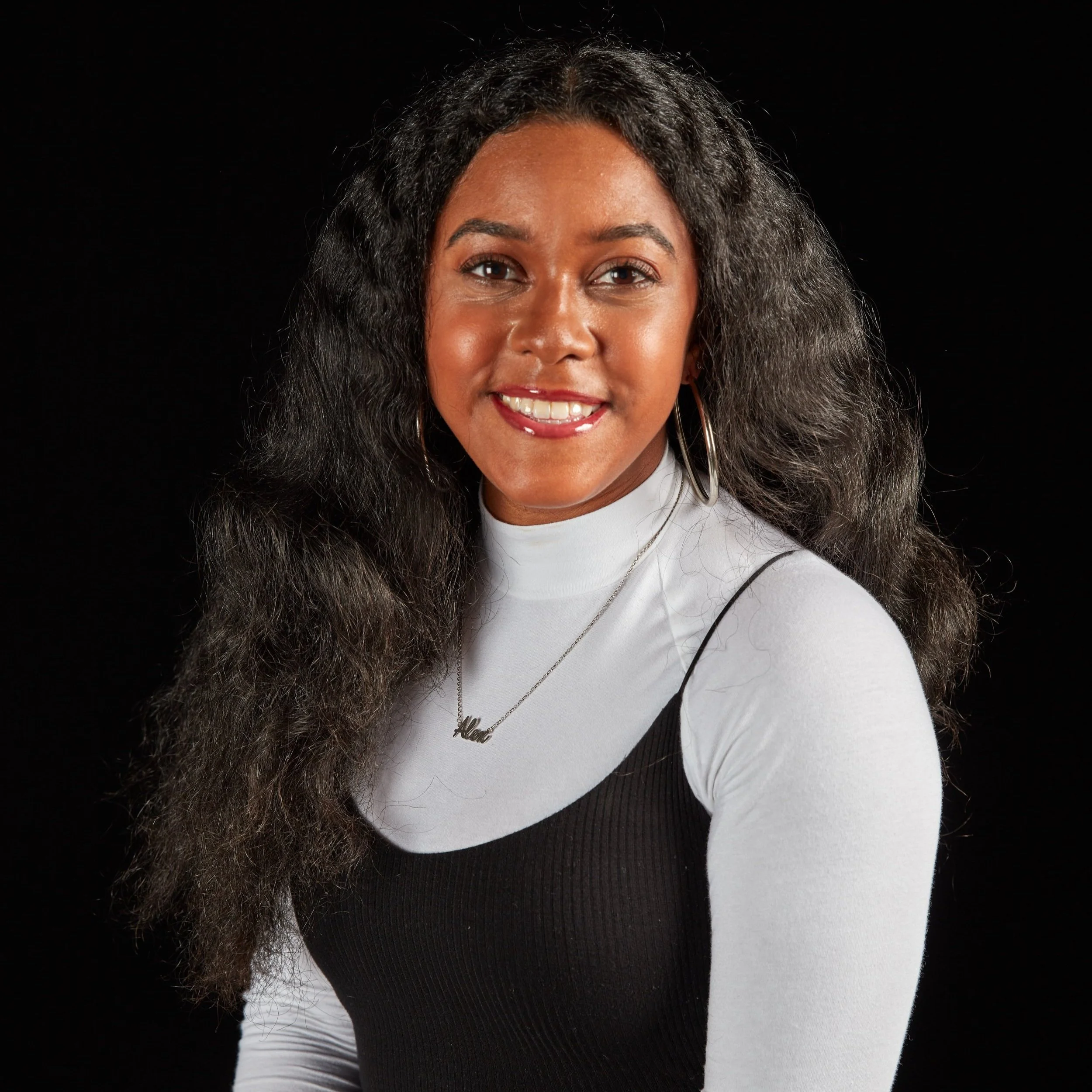 Portrait of a woman with long, wavy black hair, wearing a white turtleneck with a black dress over it, smiling, against a black background.