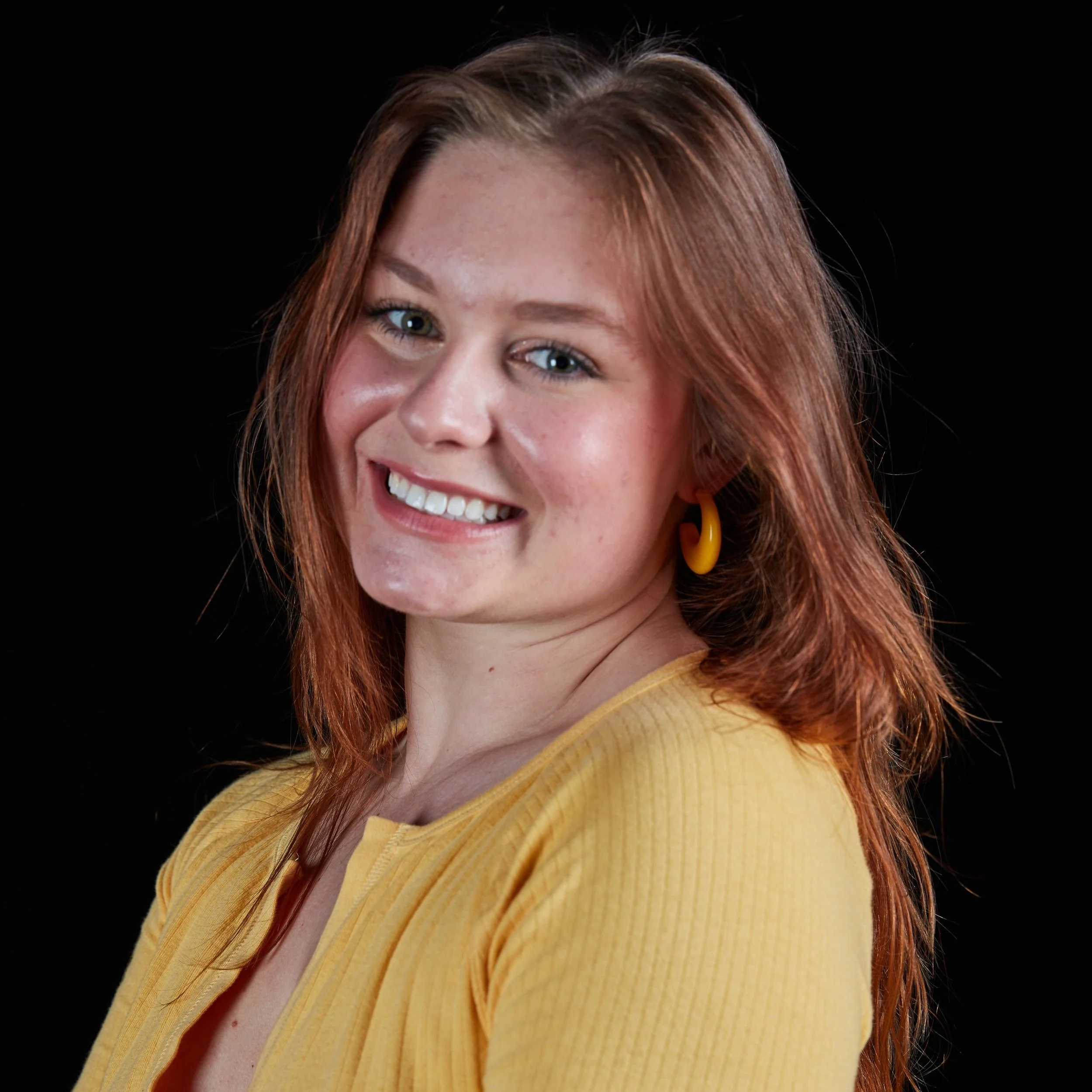 A woman with shoulder-length red hair, light skin, blue eyes, smiling, wearing yellow earrings and a yellow top, against a black background.
