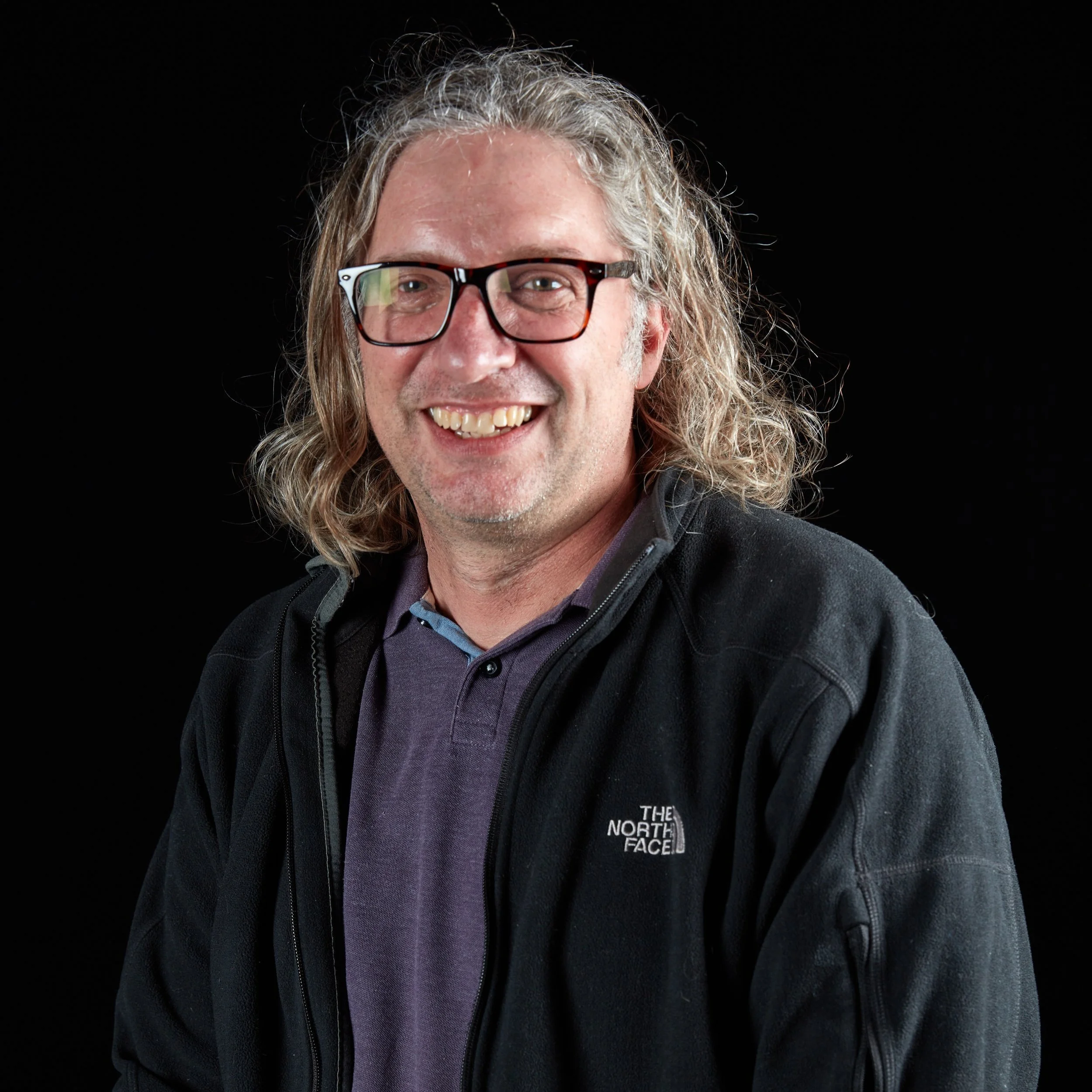 A smiling man with long, curly gray hair, wearing glasses and a black The North Face jacket against a black background.