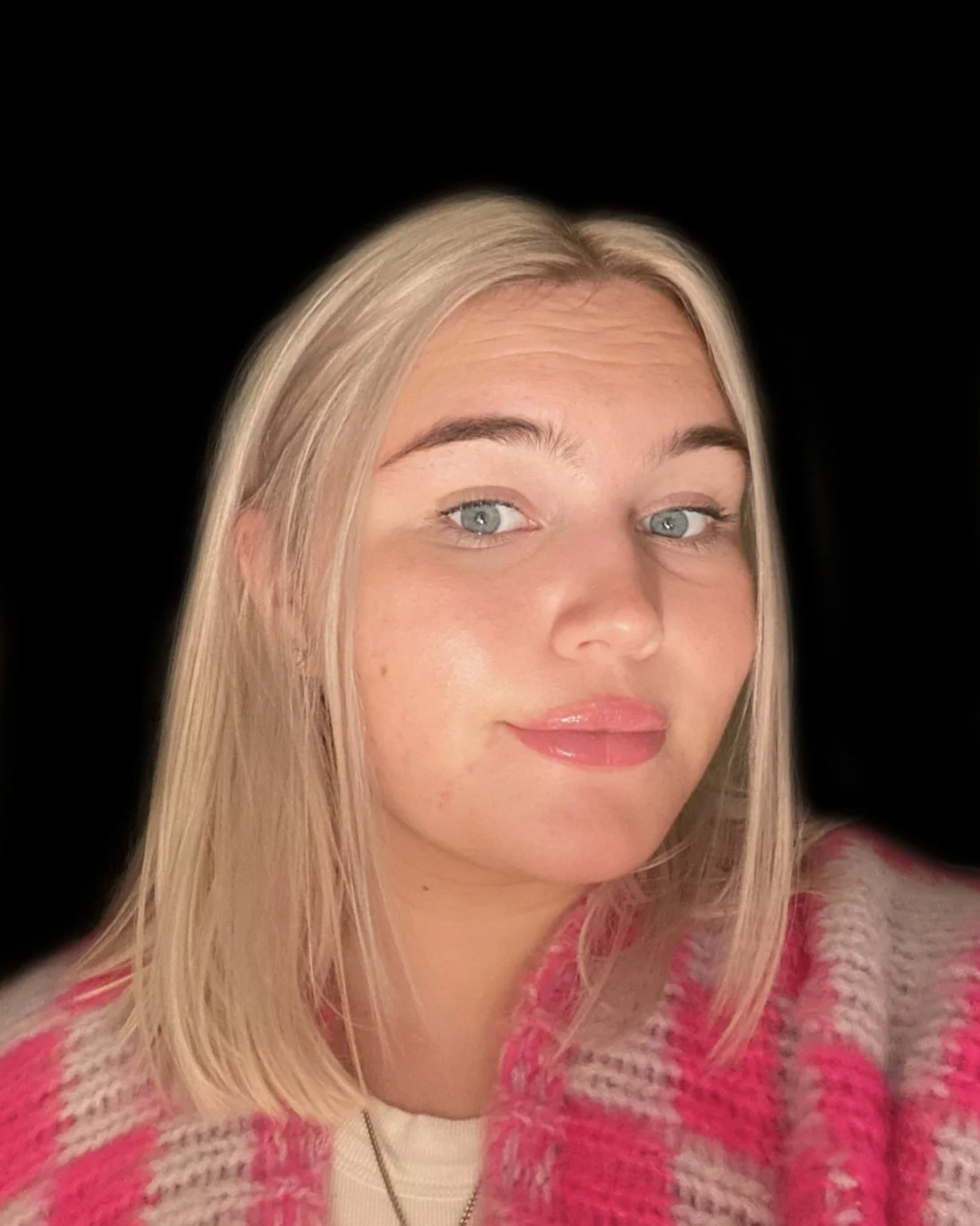 Close-up of a young woman with blonde hair, blue eyes, wearing a pink and gray plaid sweater, with a black background.