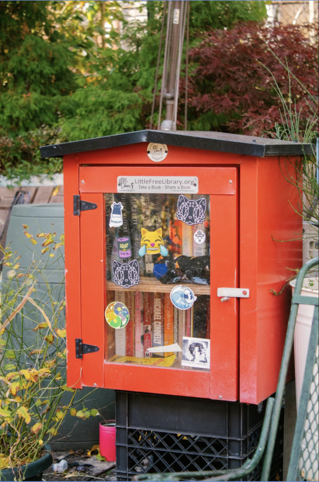 A bright red Little Free Library decorated with stickers sits among plants at Mantua’s Cat Park in West Philly