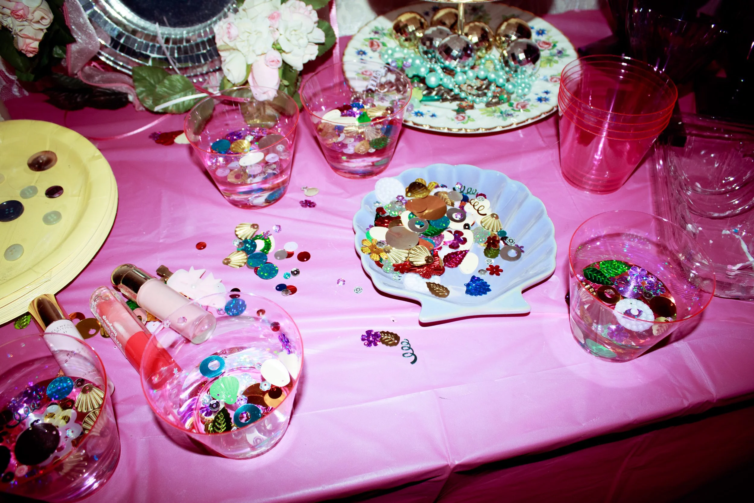  A pink Y2K-inspired craft table covered in sequins, beads, lip gloss, and coquette-style accessories arranged in pastel bowls and cups. 
