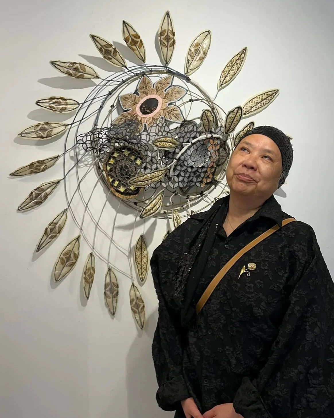 A person wearing a black patterned outfit and headscarf stands in front of a handmade wall sculpture featuring layered wire forms and floral-inspired motifs.