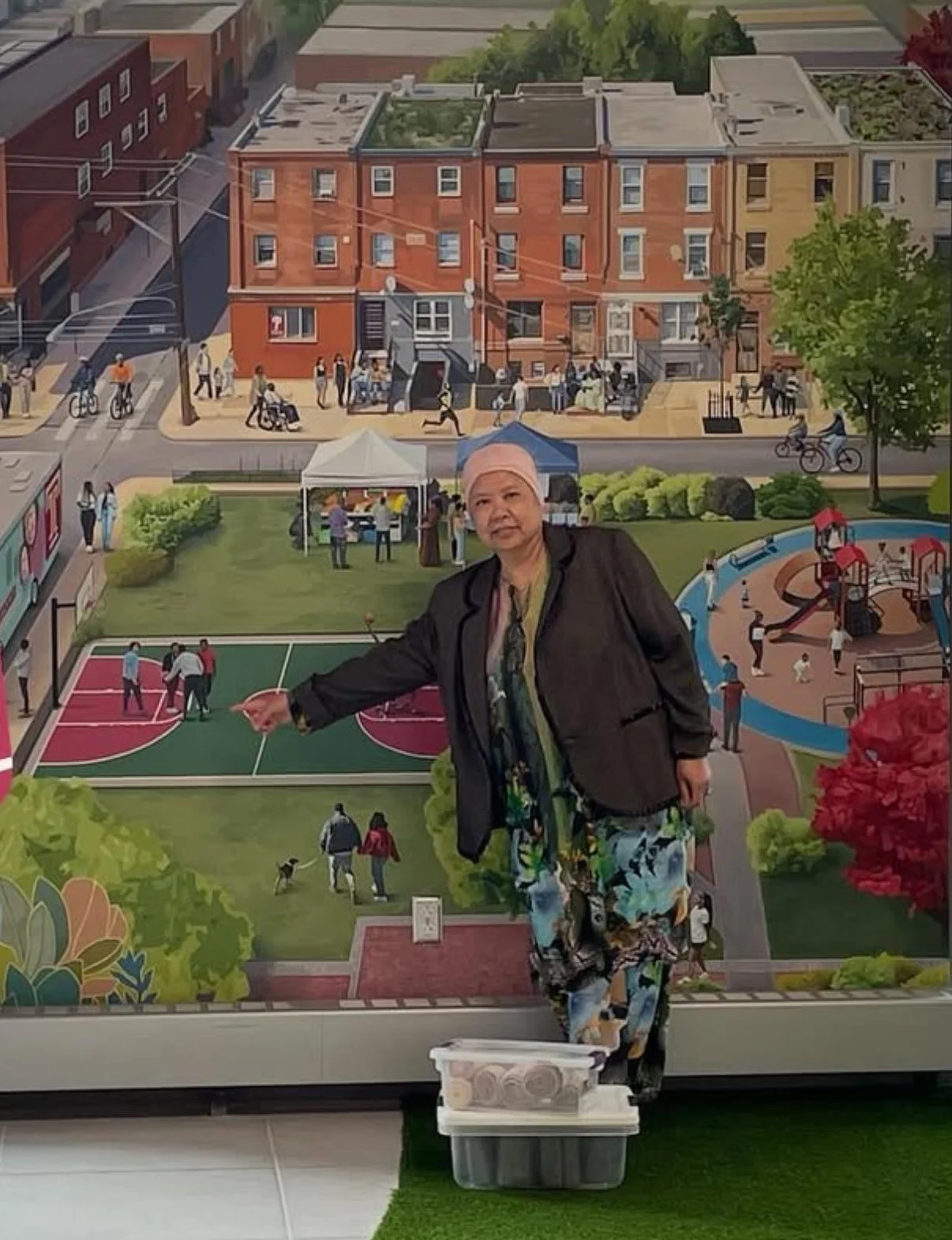 A person wearing a patterned outfit and headscarf stands in front of a large, colorful mural they created for Temple University. 