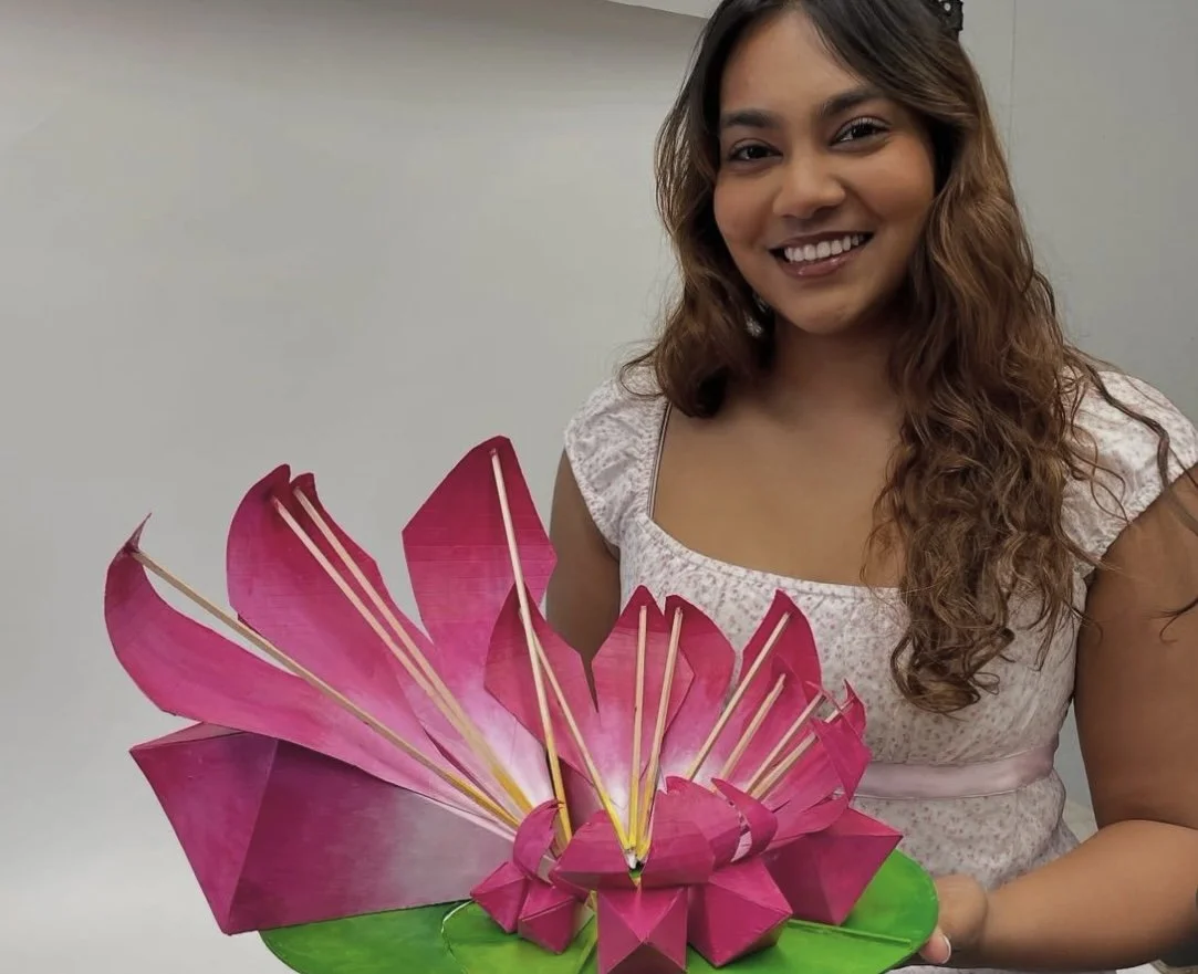 A person smiles while holding a large handmade lotus sculpture created from painted Bristol board and wooden sticks, designed using the Fibonacci sequence for a Design 3 final project.