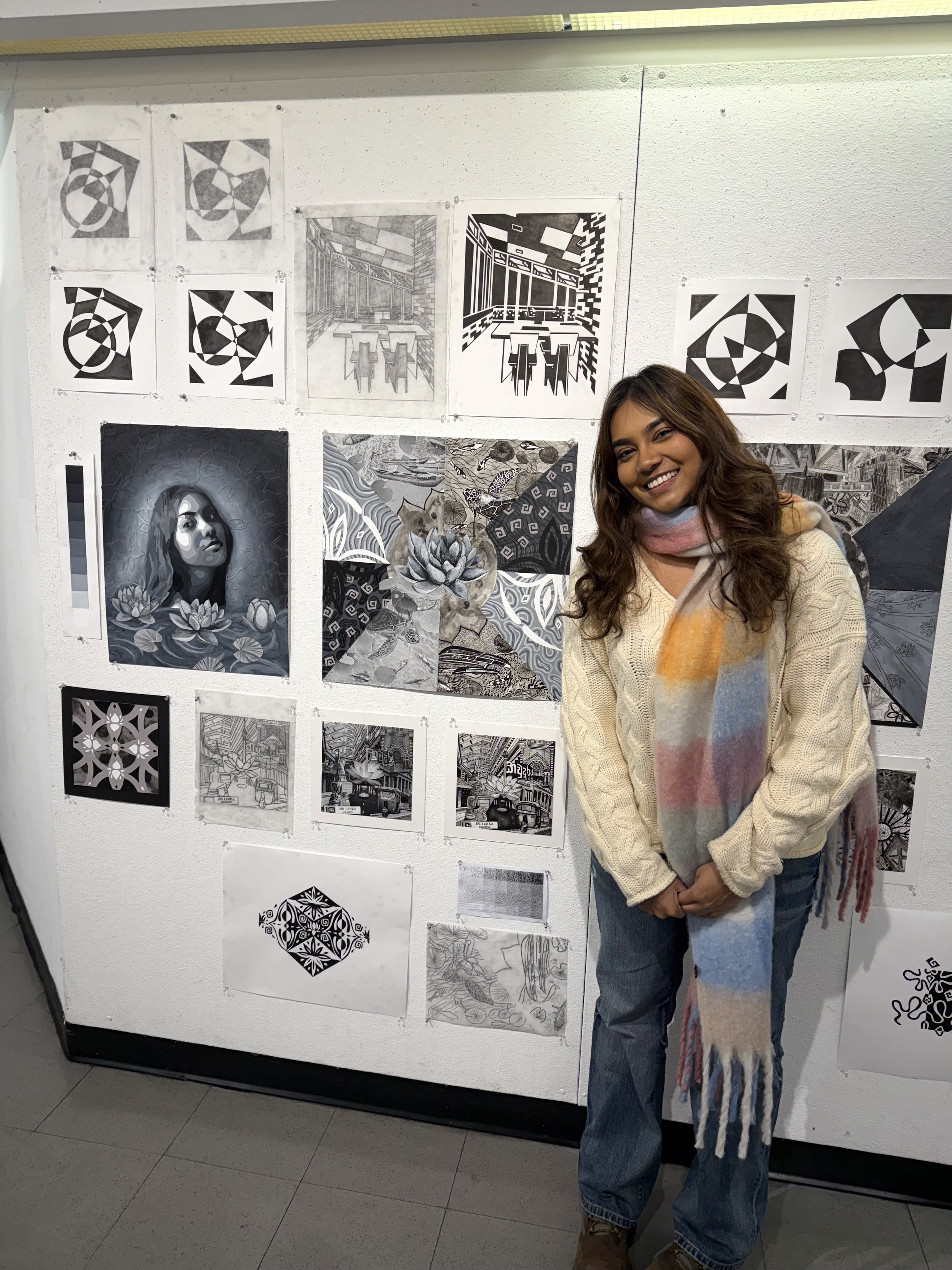 A person stands smiling next to a wall covered with their Design 1 portfolio, including geometric drawings, perspective sketches, mixed-media pieces