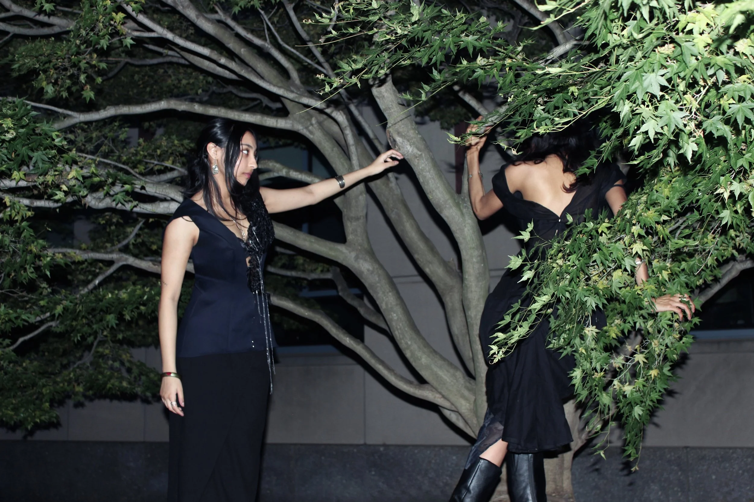An outdoor nighttime editorial photoshoot shows two people posing among tree branches while wearing black fashion designs. On the left, one person wears a structured black vest made from recycled.