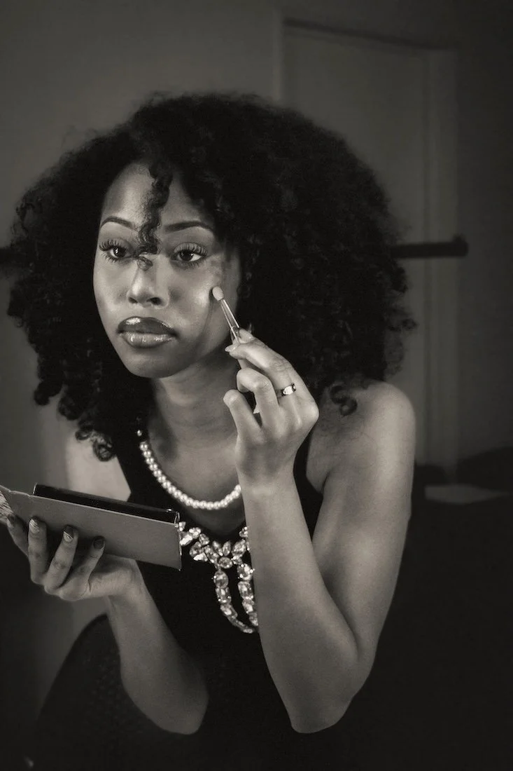 A woman with voluminous curly hair applies makeup with a brush while looking into a dressing room mirror. She wears a black dress with jeweled accents and a pearl necklace. 