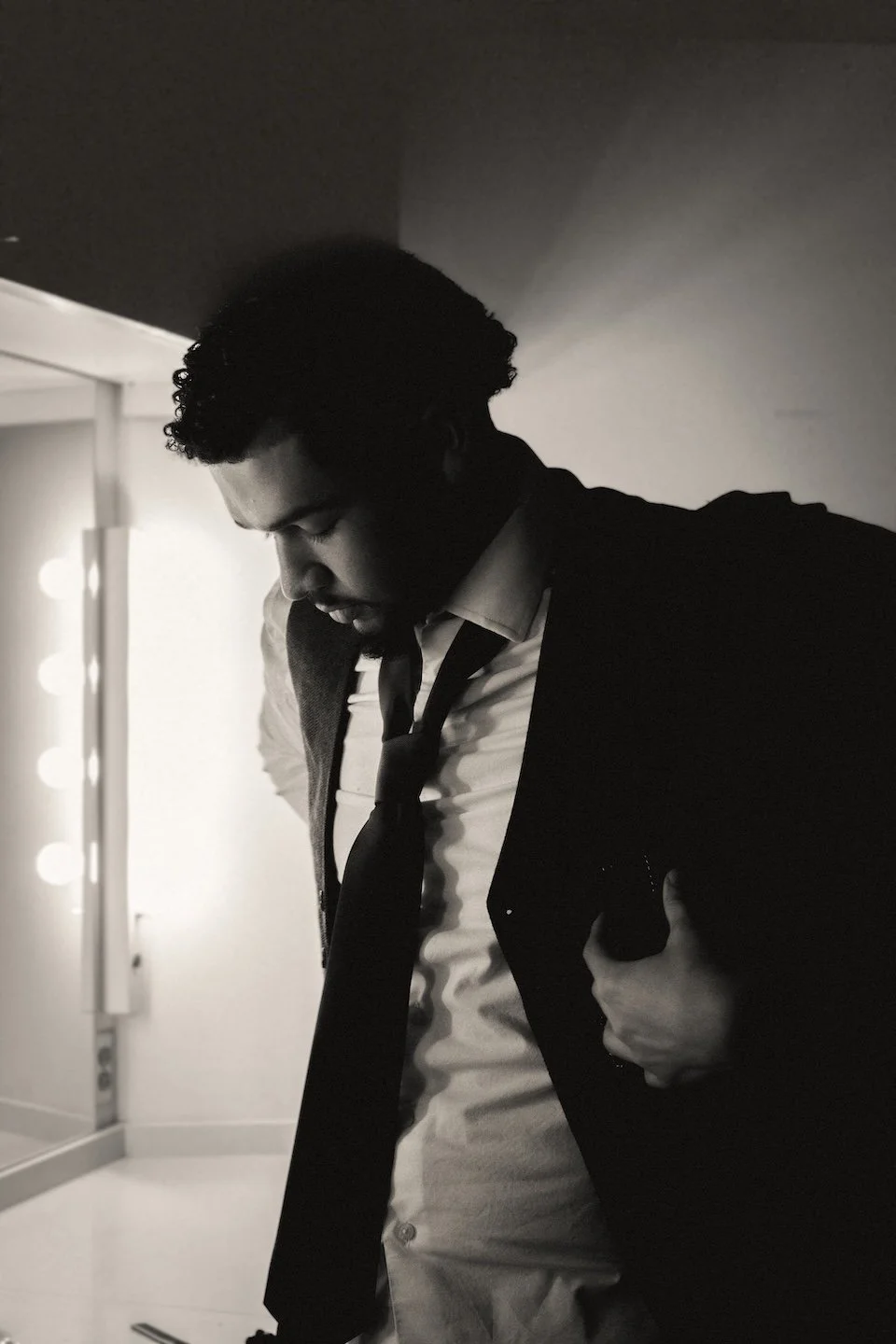 A man with short curly hair adjusts his suit jacket in a softly lit dressing room, wearing a loosened tie and button-down shirt. 
