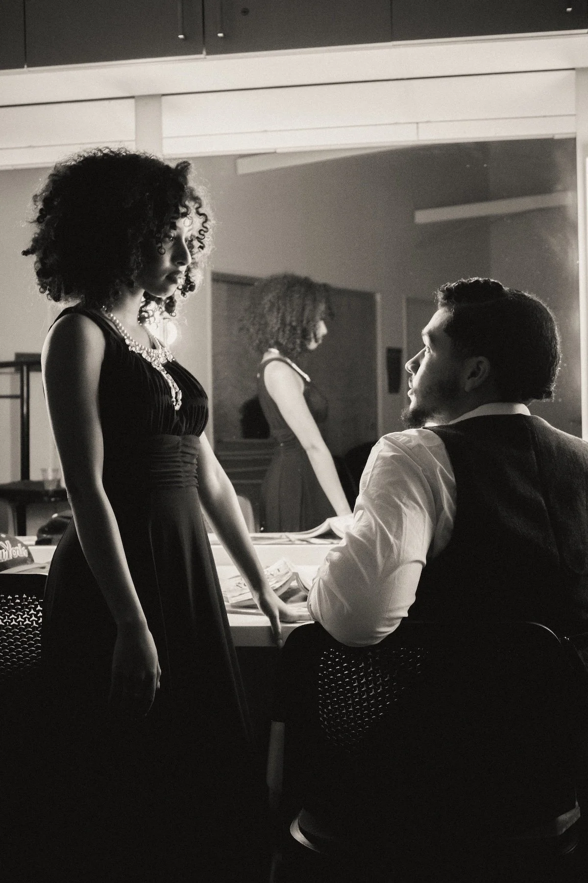 A woman with curly hair in a long black dress stands above a seated man in a dressing room lit by mirror lights, both appearing to be in conversation before a performance. 
