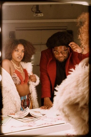  A woman with curly hair, wearing a fur coat and a bold 70s-inspired outfit, stands beside a man with an afro, both looking into a brightly lit mirro