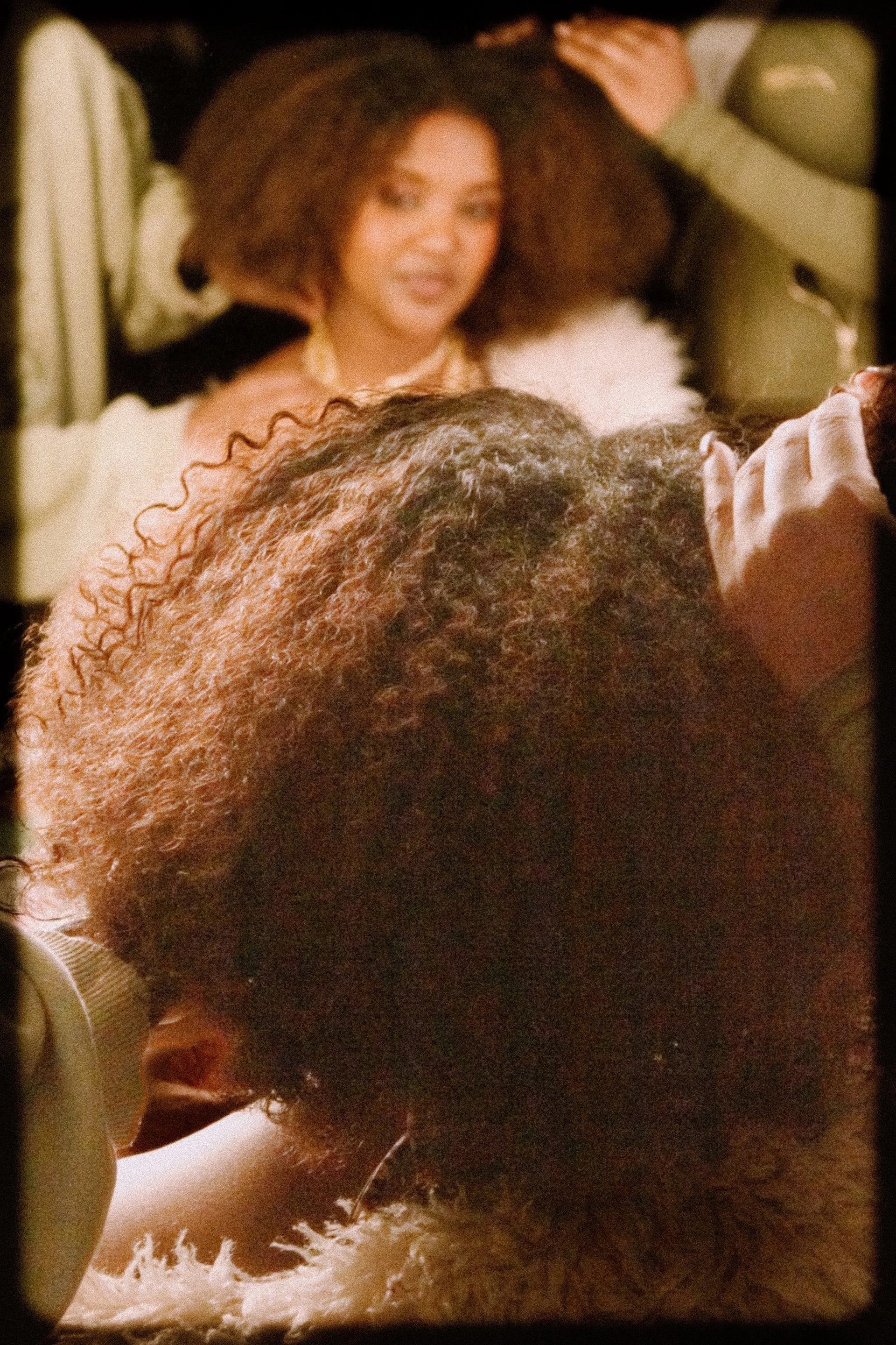 A woman with voluminous curly hair sits in front of a mirror while someone styles her hair, her reflection softly in focus and framed by warm, vintage lighting. 