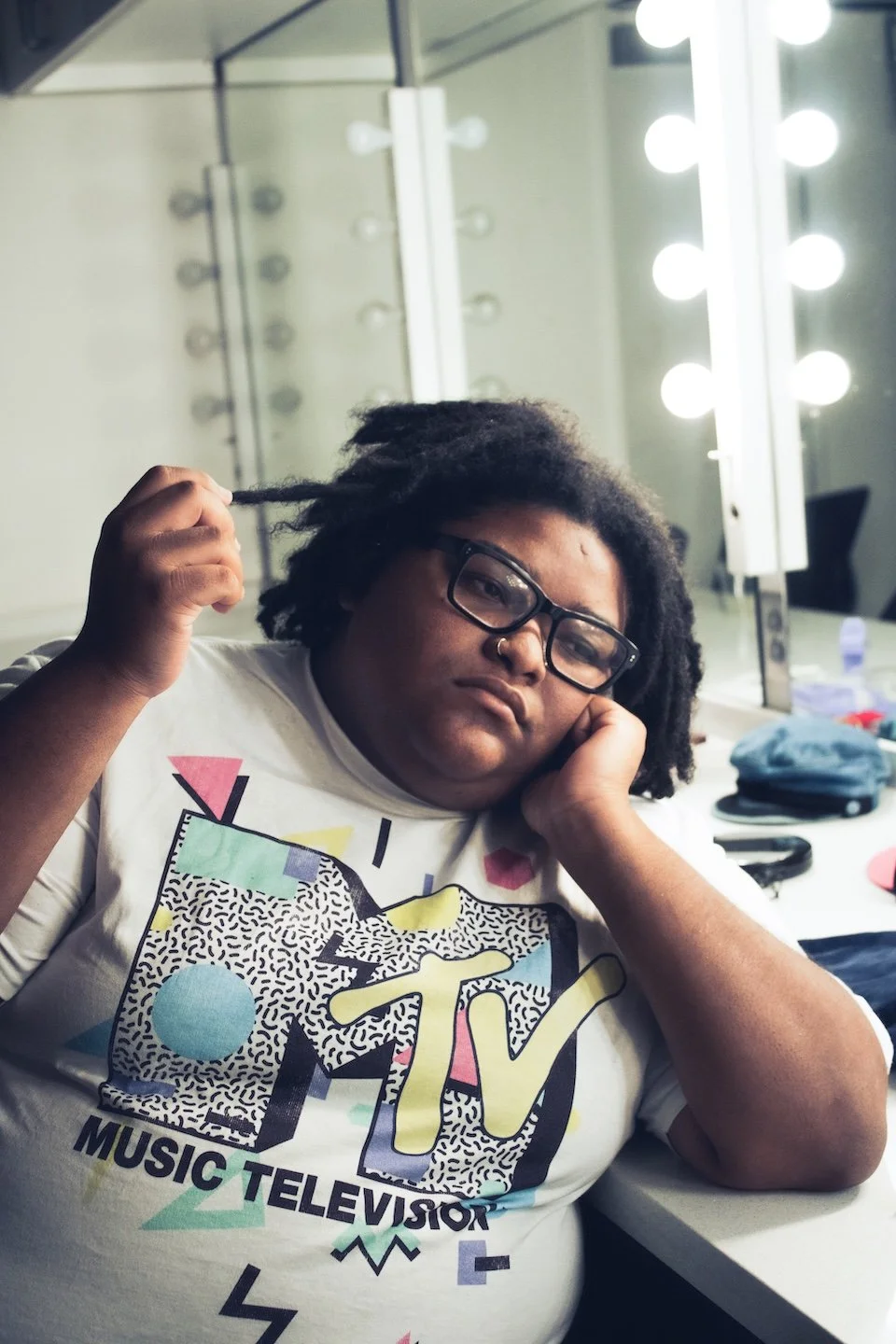 A person wearing an MTV graphic T-shirt sits in a dressing room, gently holding one of their locs while resting their head on their hand beneath bright vanity lights.
