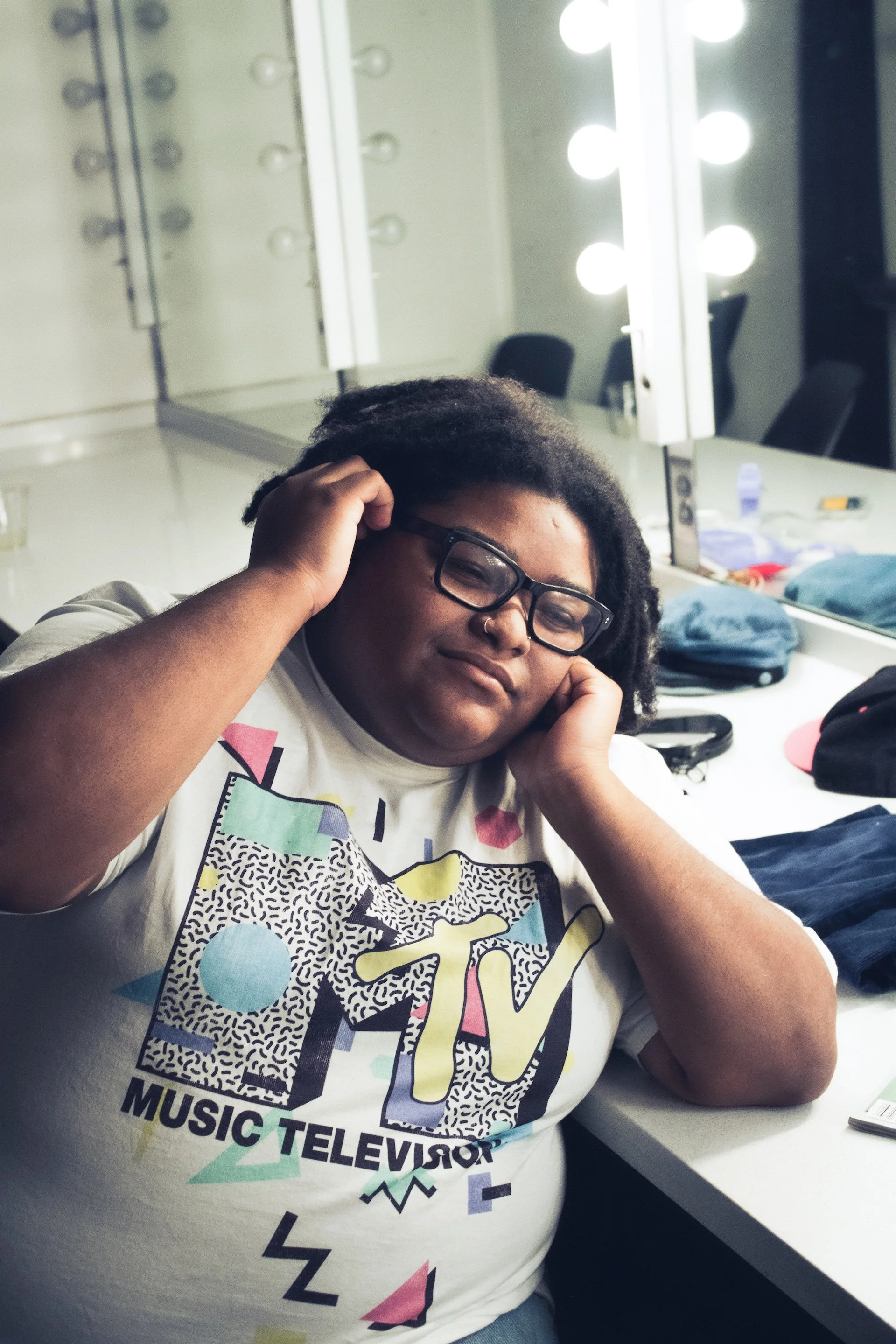 A person wearing an MTV graphic T-shirt sits in a dressing room, gently touching their locs while resting their head on their hand longingly beneath bright vanity lights. 