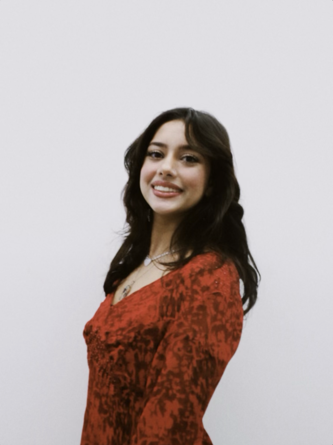 A young woman with dark hair wearing a red patterned dress and a necklace, standing against a plain light-colored background, smiling at the camera.